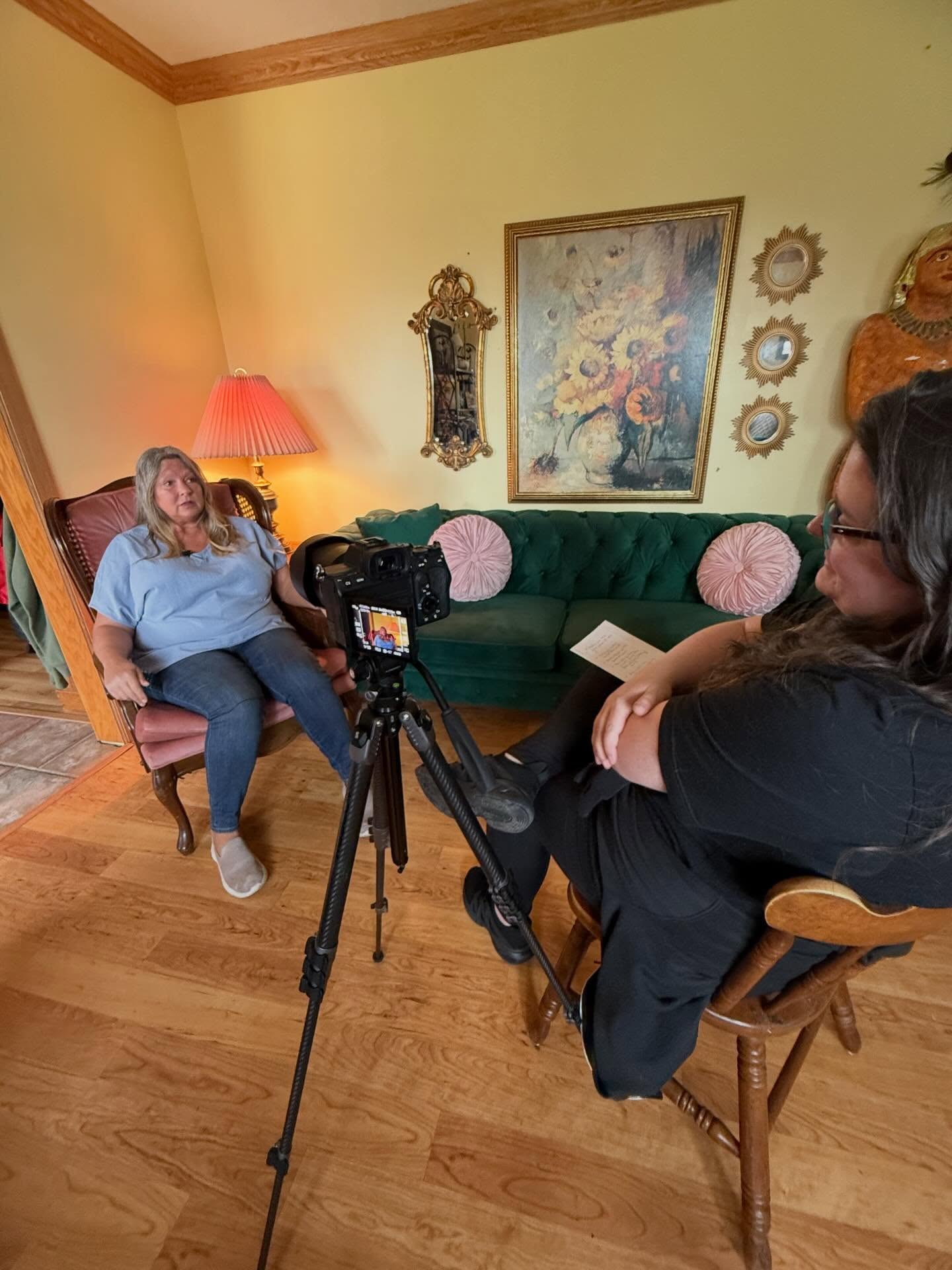 Woman sitting on a pink chair being interviewed by another woman with a camera, in a room with artwork and a green sofa.