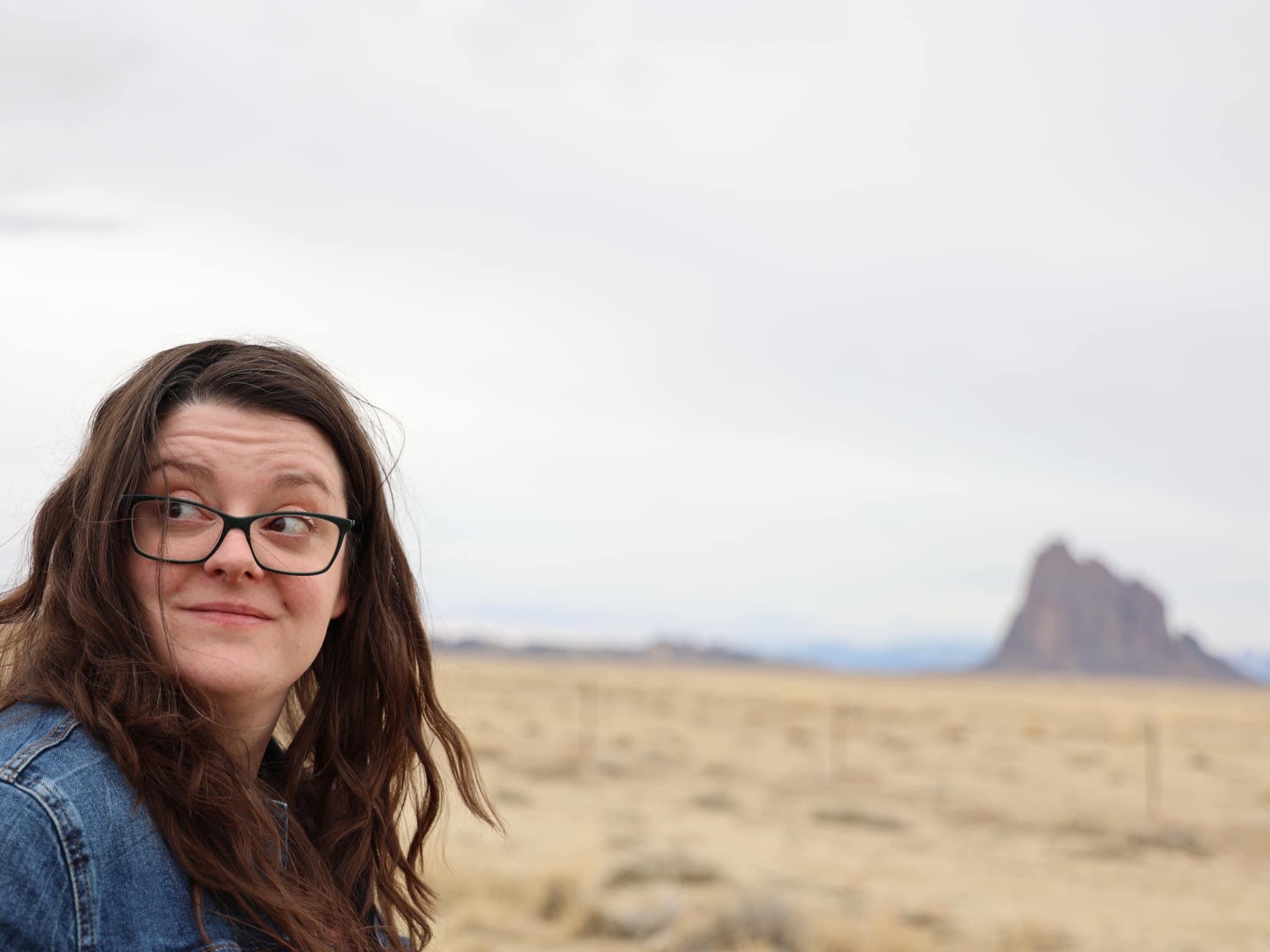 Woman with glasses and wavy hair outdoors in a desert landscape with a rock formation in the background.
