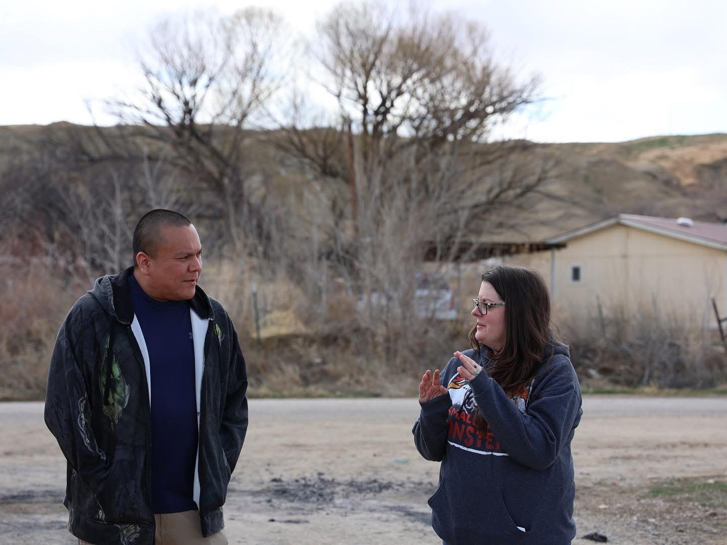 Two people standing outdoors, talking, with trees and a building in the background.