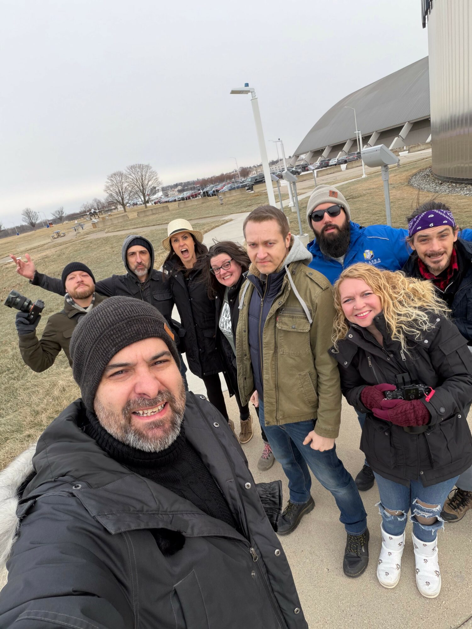 Group of eight people outdoors, smiling, standing close together, some with arms around each other, in front of a modern building and cloudy sky.