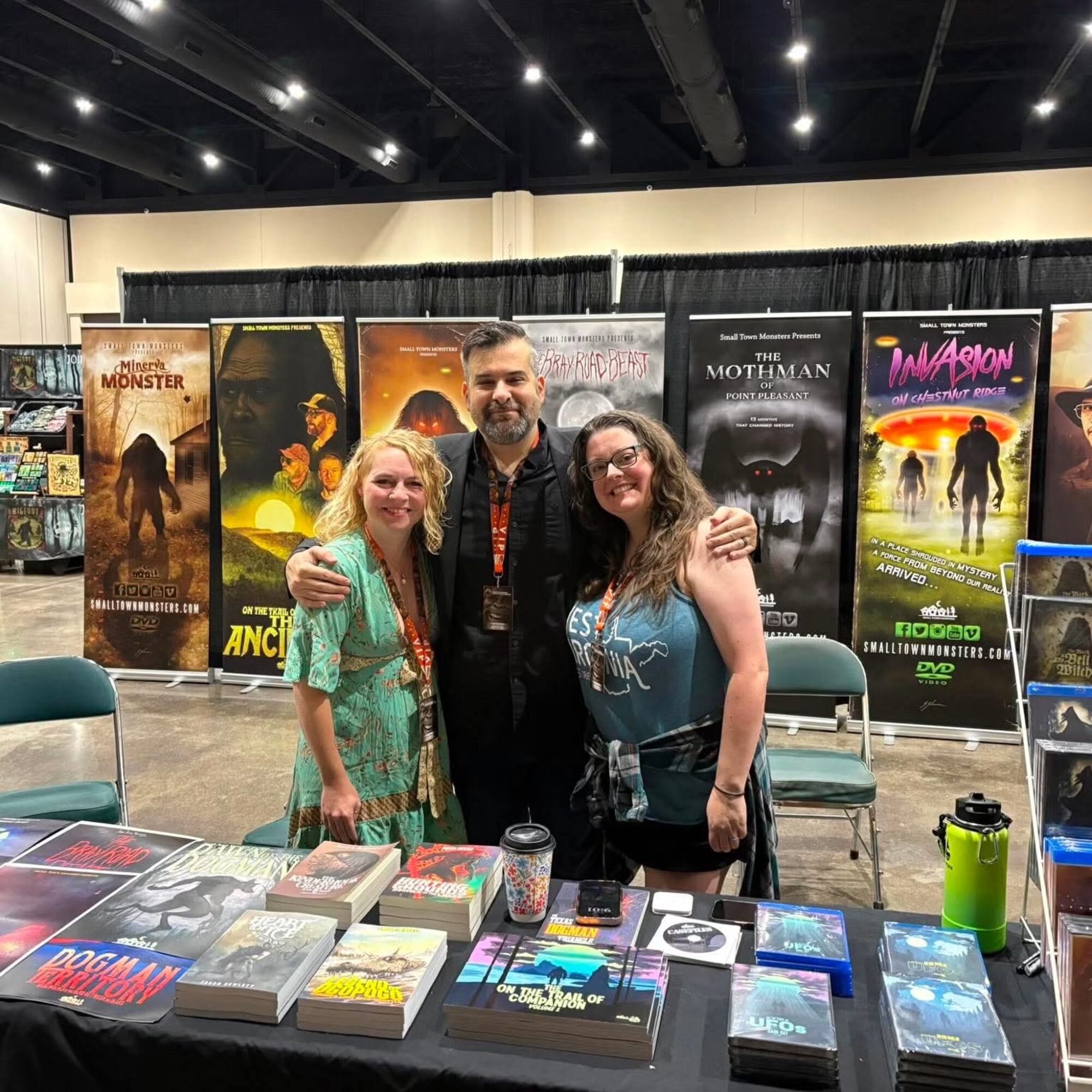 Three people standing behind a table with books and merchandise at a convention, with posters in the background.