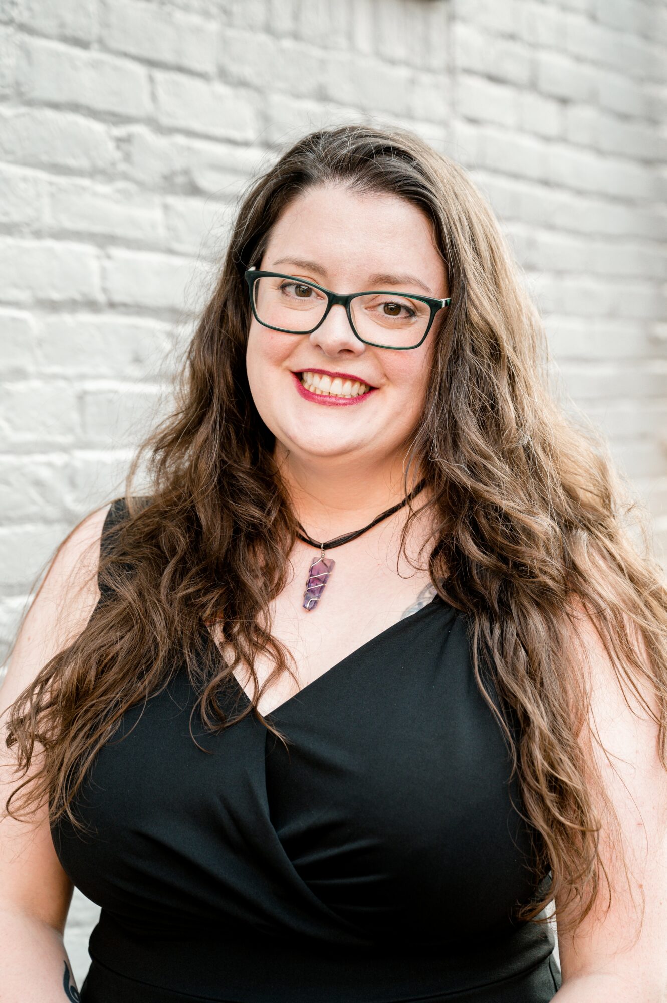 Woman with long wavy hair, glasses, and a black top, smiling in front of a brick wall.