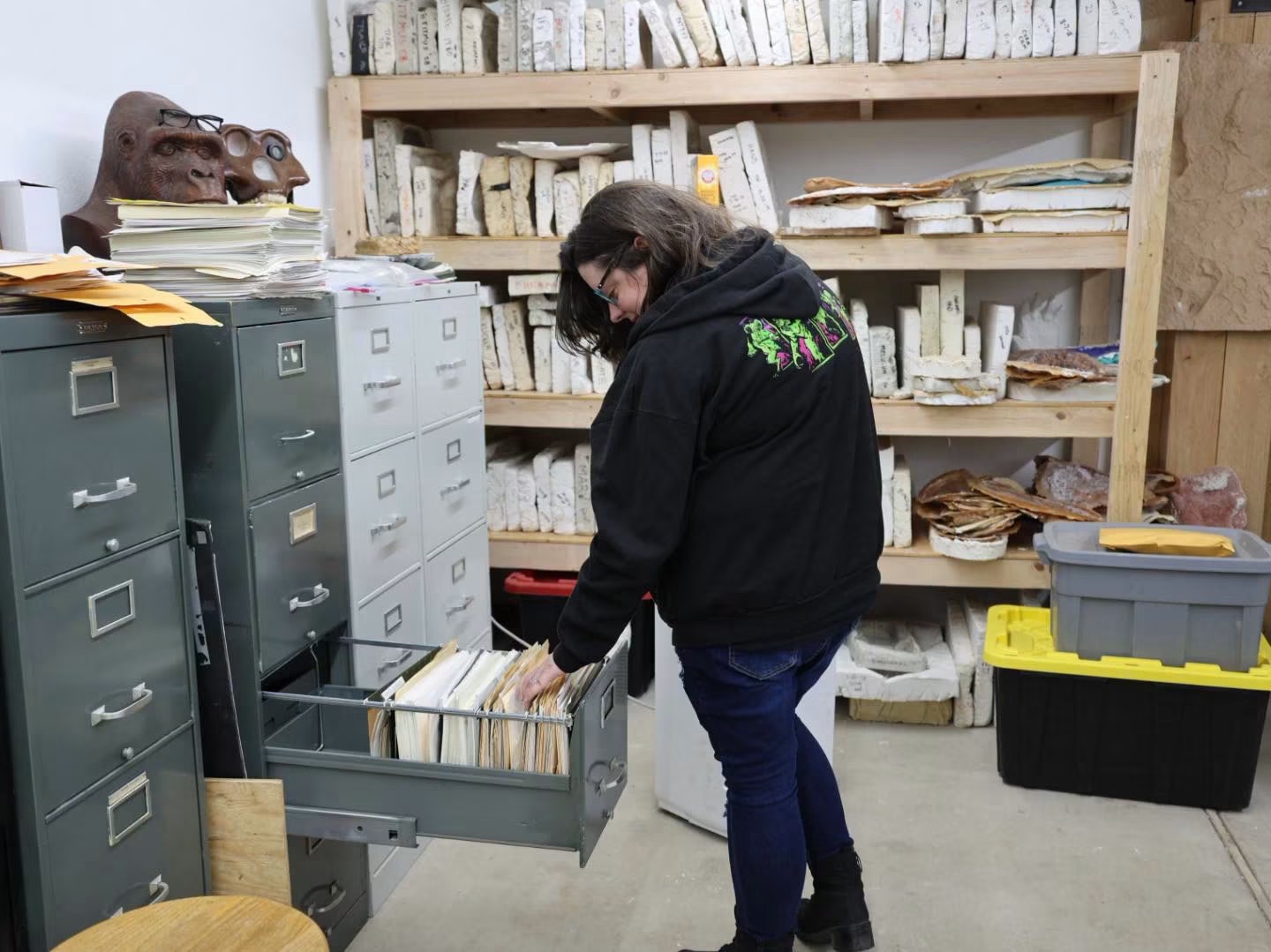 Person with dark hair and glasses, wearing a black hoodie, searches through a filing cabinet in a storage room.