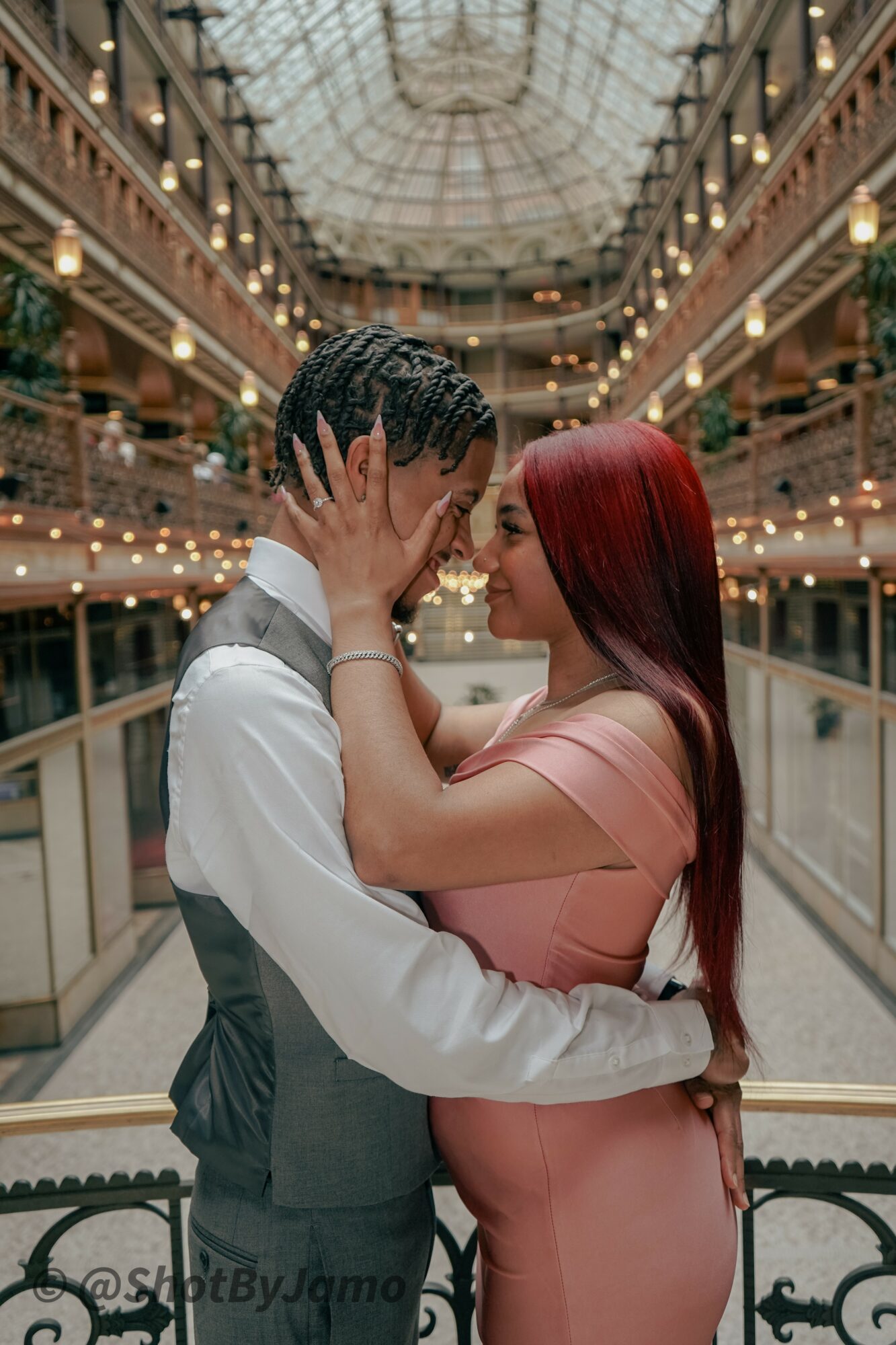 A couple stands close in a grand, ornate indoor setting with a glass ceiling, holding each other's faces and gazing at each other.