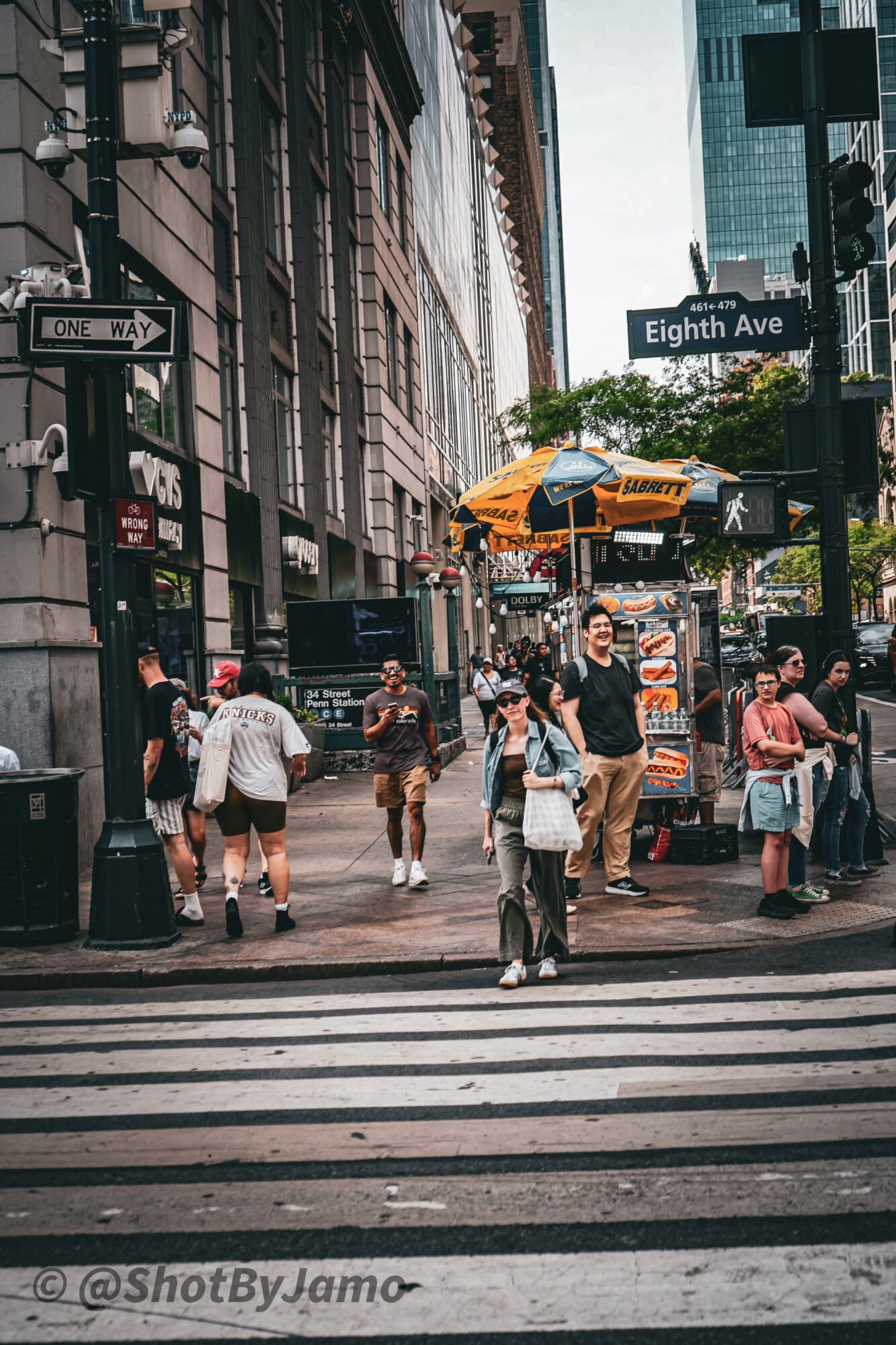 People crossing a busy city street at a crosswalk with tall buildings and street signs, including Eight Avenue, in the background.