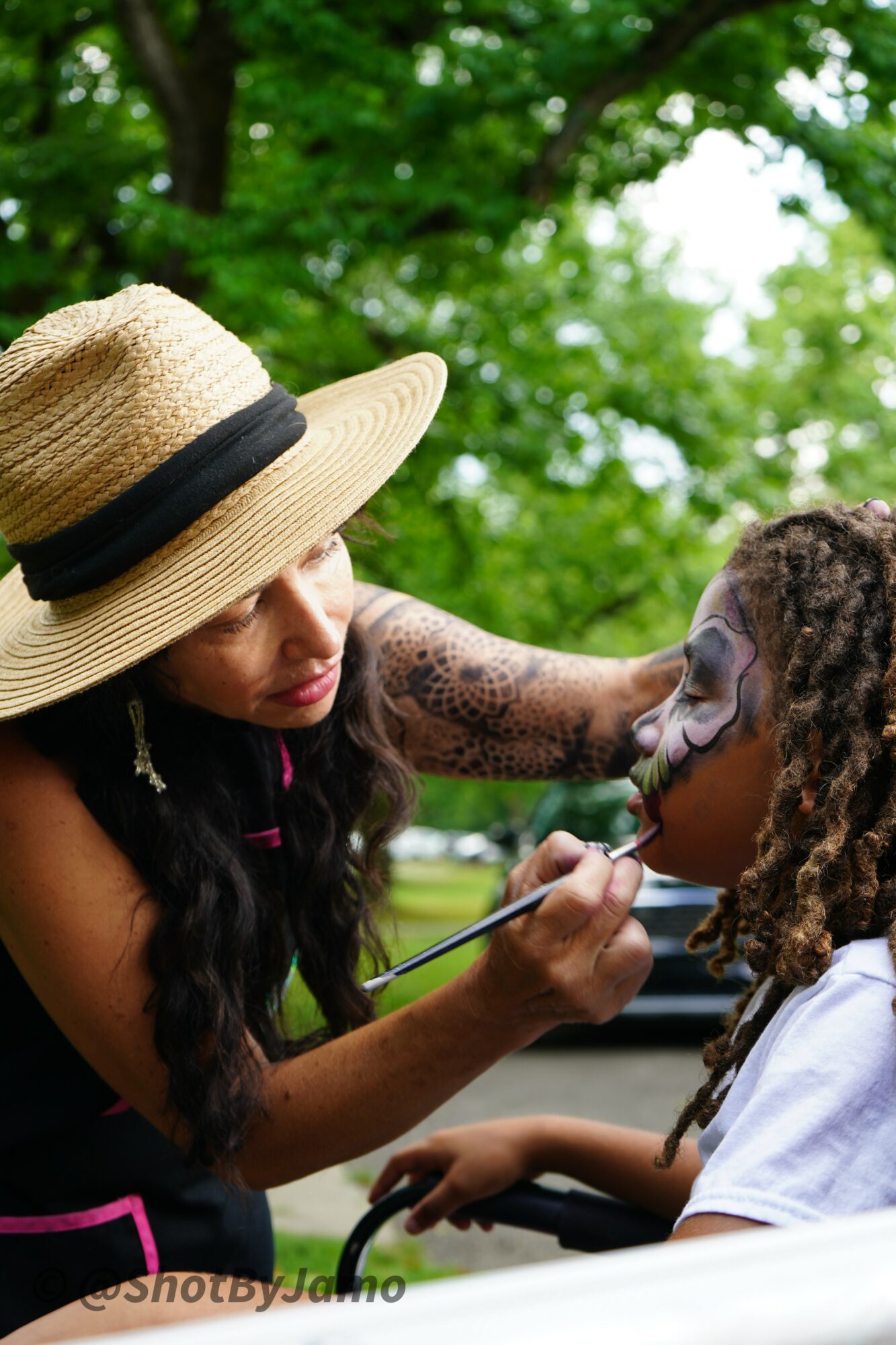Woman applying face paint to a child's face outdoors with green trees in background.