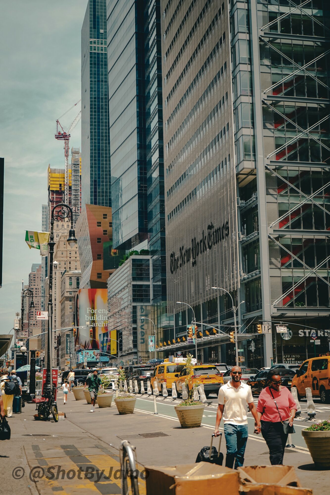 City street with tall buildings, yellow taxis, pedestrians, and construction cranes, under cloudy sky.
