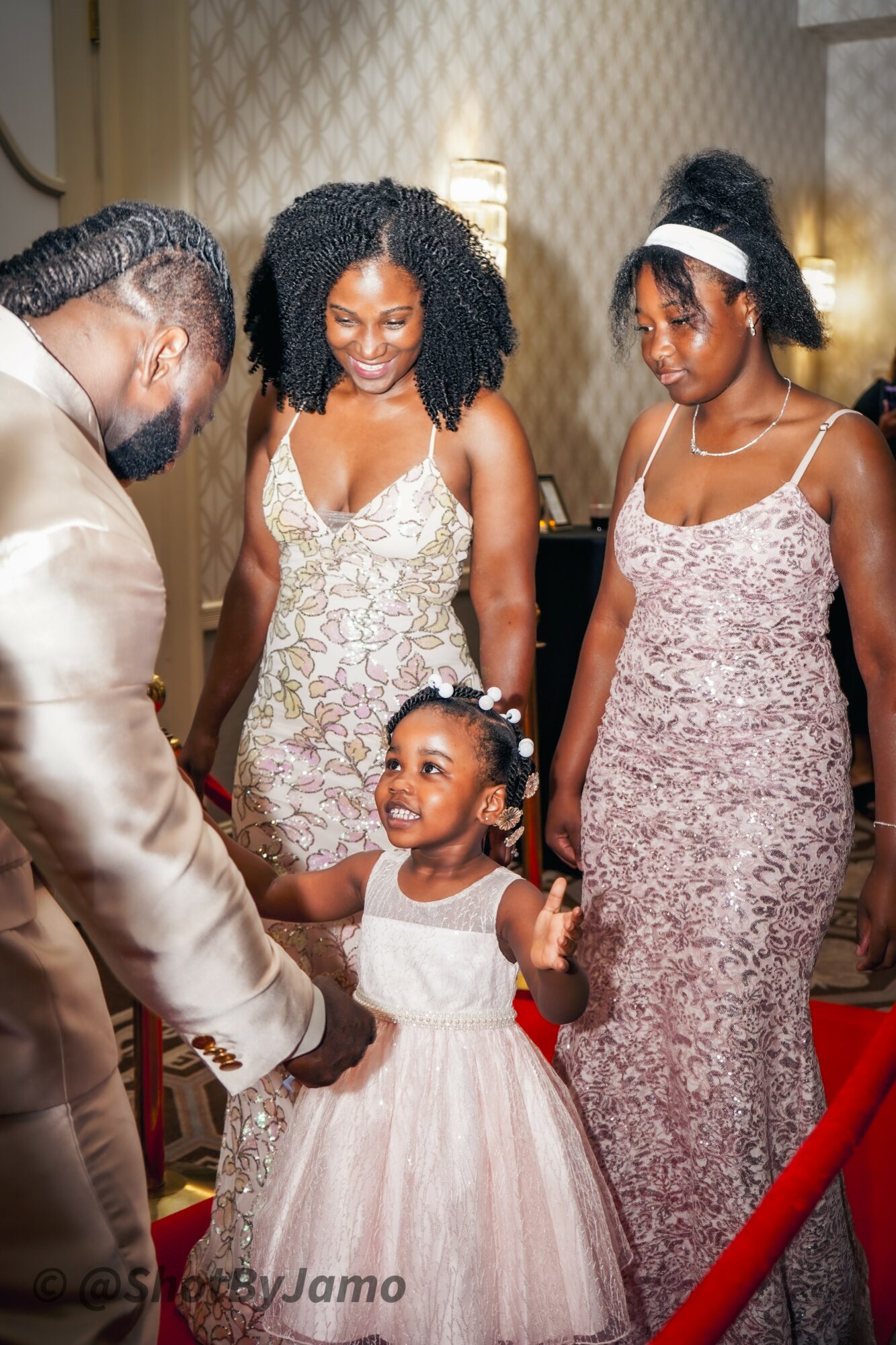 Four people, including a young girl in a white dress, smiling and holding hands at an indoor event.
