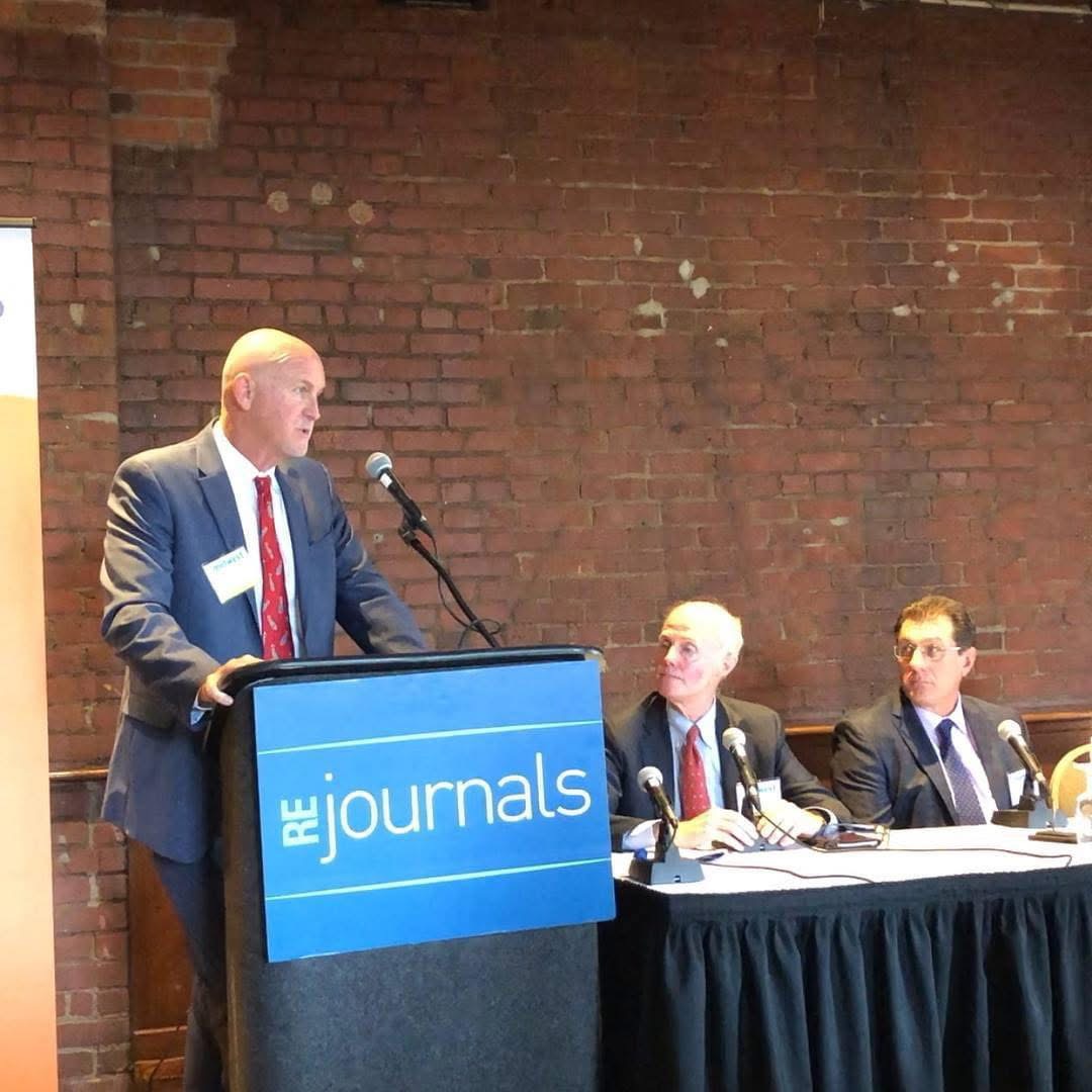 Man in suit speaking at a podium with a microphone, two men seated at a table, brick wall background.