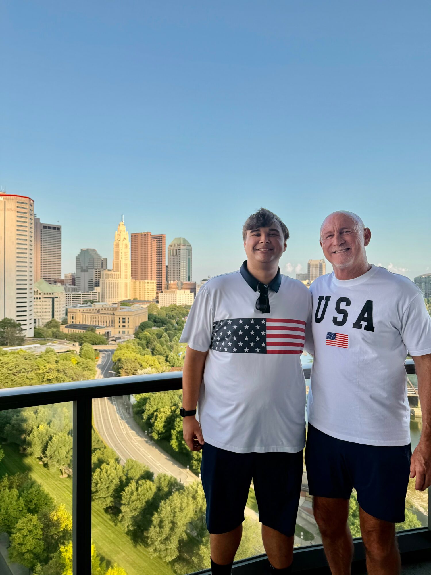 Two people stand on a balcony with a city skyline and green park in the background, smiling at the camera.