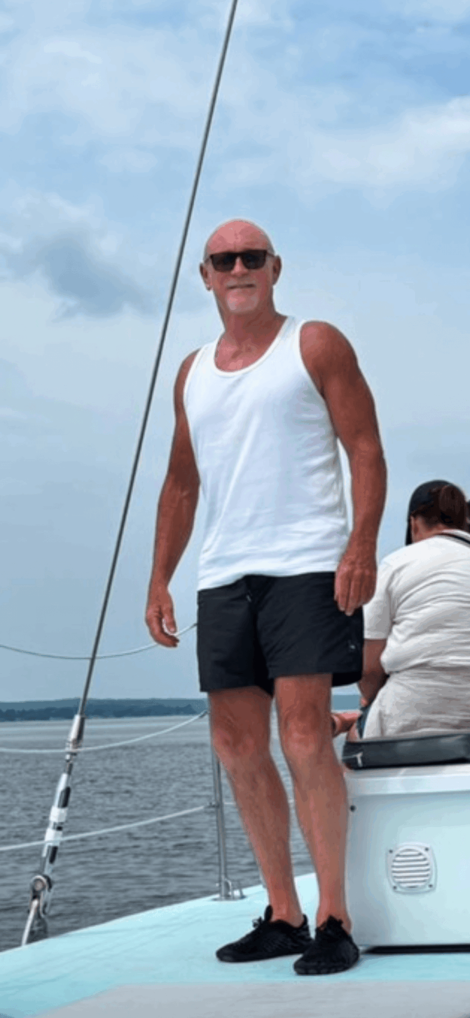 Man in white tank top and black shorts standing on boat, with water and sky in background.