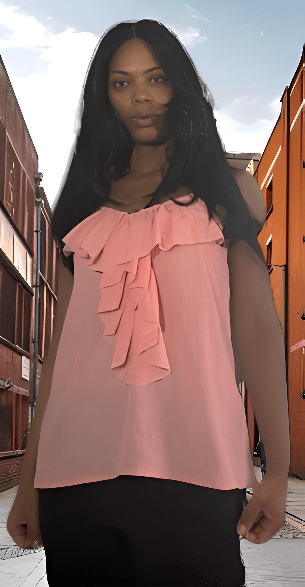 Woman with long dark hair wearing a pink ruffled top standing outdoors with buildings in background.