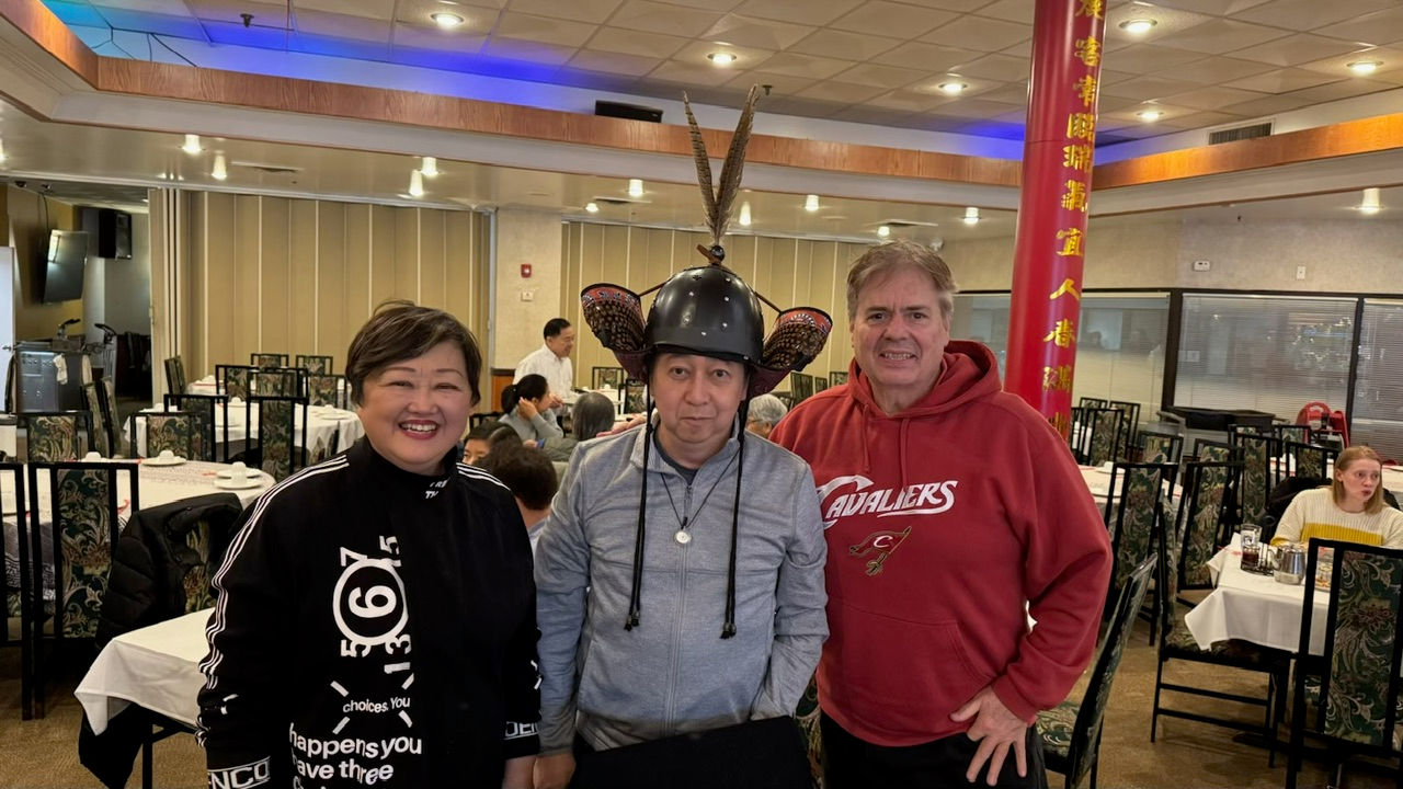 Three people standing in a restaurant, one wearing a helmet with antennae, two smiling, tables and other diners in background.