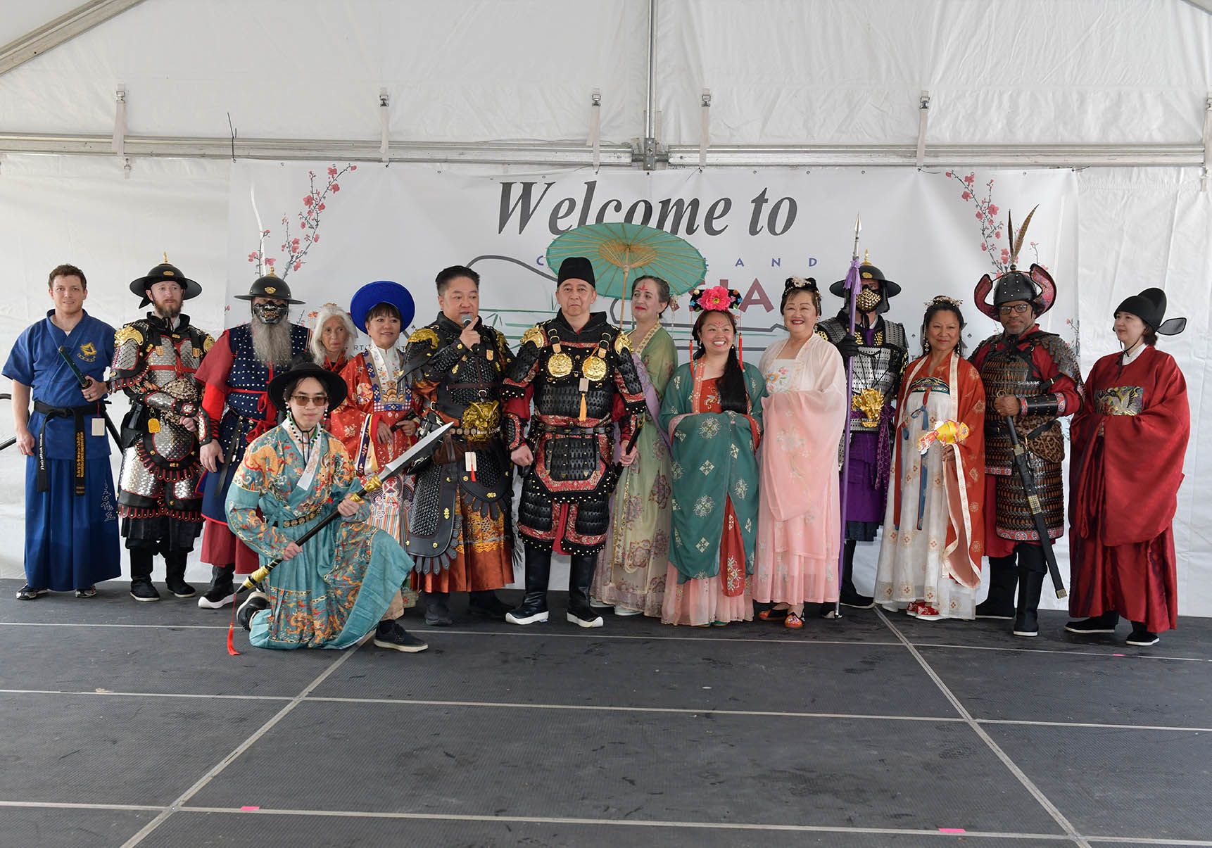 Group of people in traditional costumes standing on stage with a 'Welcome to' banner behind them.