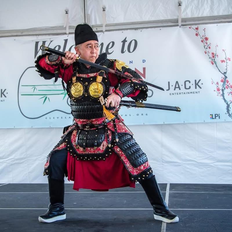 Person in traditional costume holding a sword in a martial arts stance on stage, with a banner in the background.