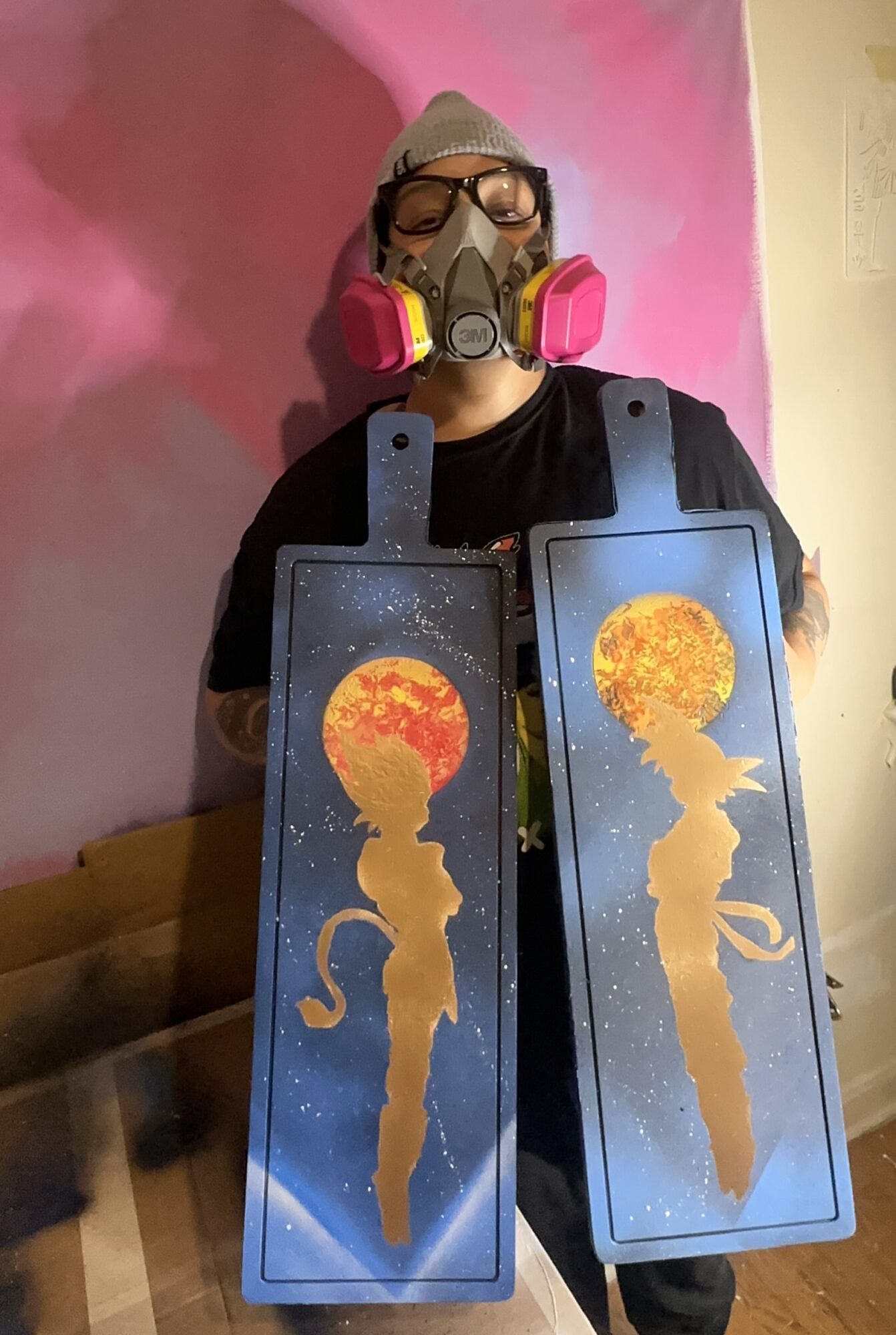 Person wearing a mask and glasses holding two rectangular signs with a silhouette of a woman and a fiery explosion, against a pink background.