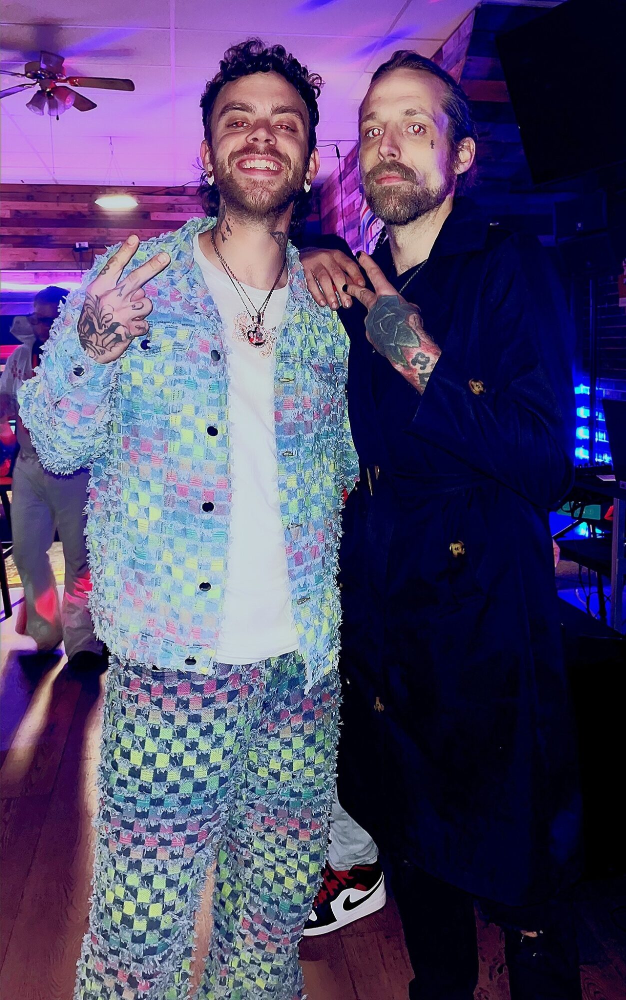 Two men pose together in a colorful indoor setting, one smiling and making a peace sign, the other with a serious expression.