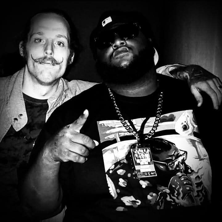 Two men pose together, one with a mustache and the other wearing sunglasses and a cap, in black and white.