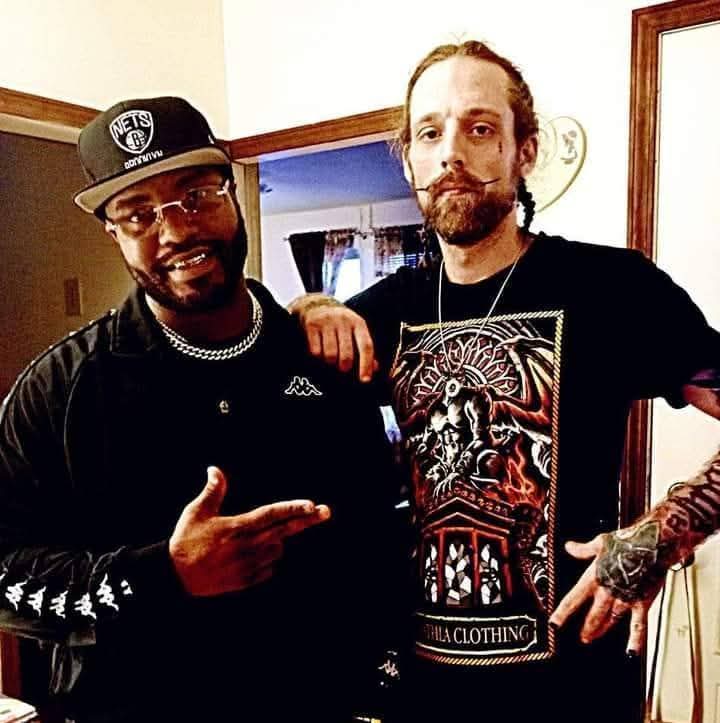 Two men standing indoors, one wearing a cap and glasses, the other with a beard and tattoos, both smiling.