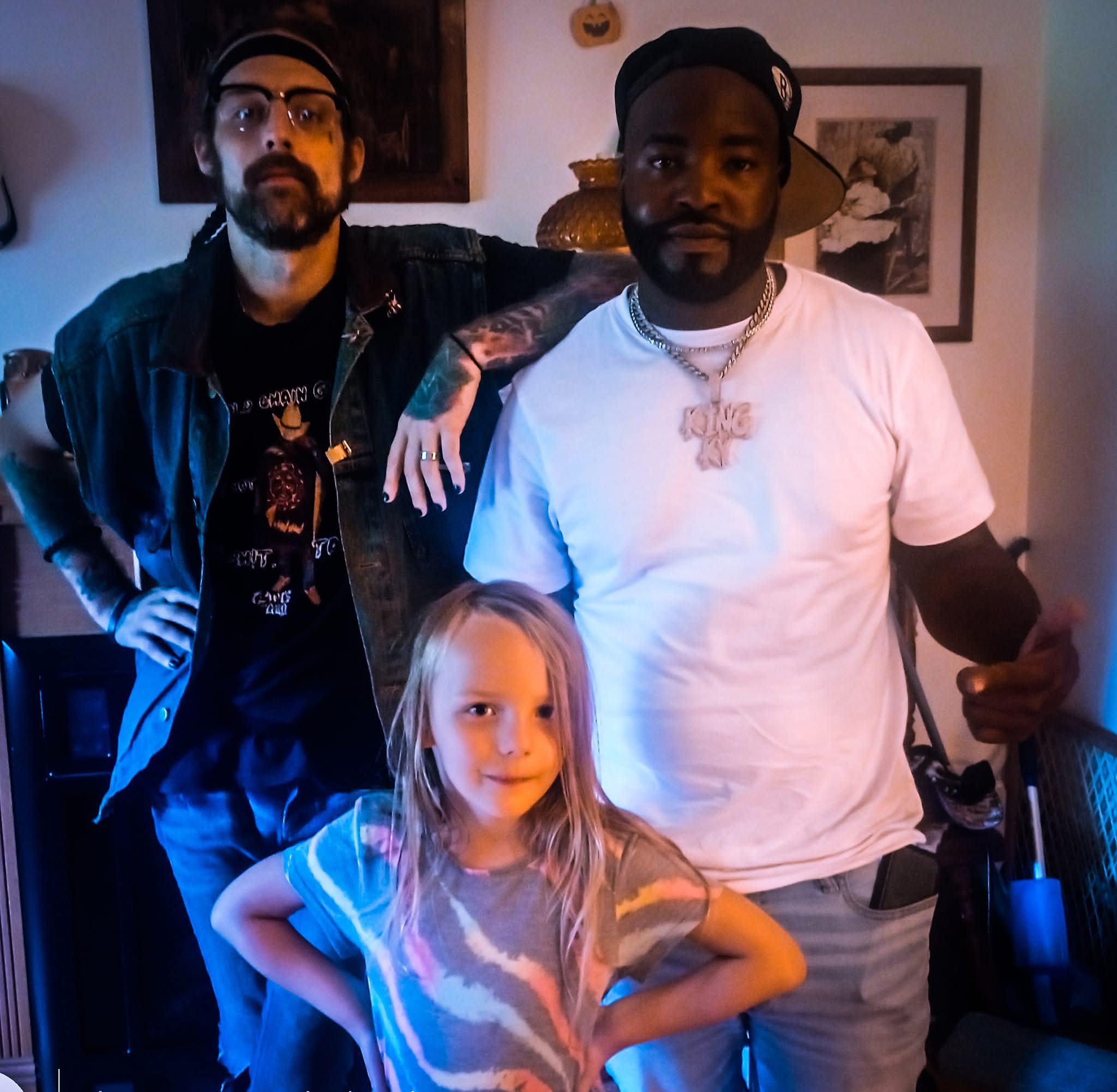 Two men and a girl pose indoors, one man with glasses and tattoos, the other in a white t-shirt, girl in front.