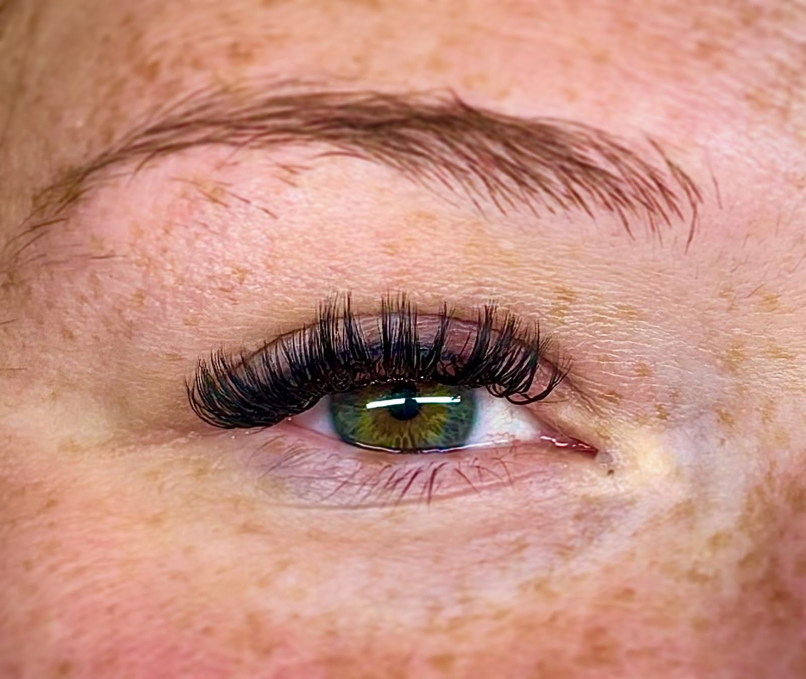 Close-up of a person's eye with long eyelashes and an eyebrow, showing green eye color and freckles.