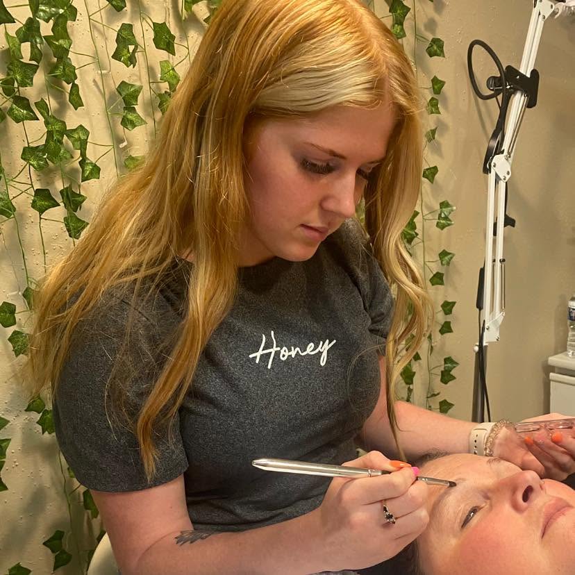 Woman with long blonde hair applying makeup to a person's face, surrounded by green leafy decor.