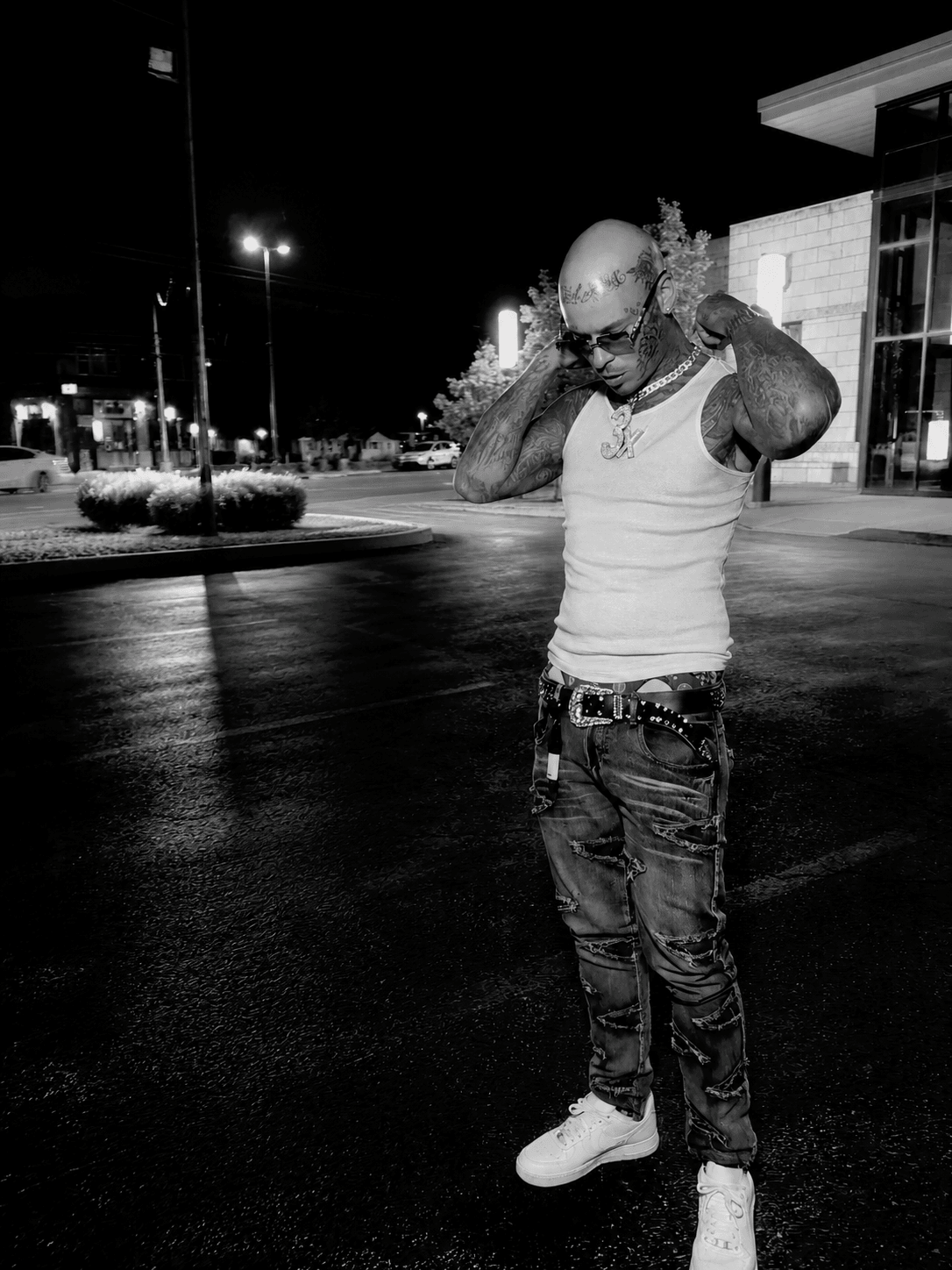 Man with sunglasses and tattoos adjusting his shirt outside at night, illuminated by streetlights, in front of a building.
