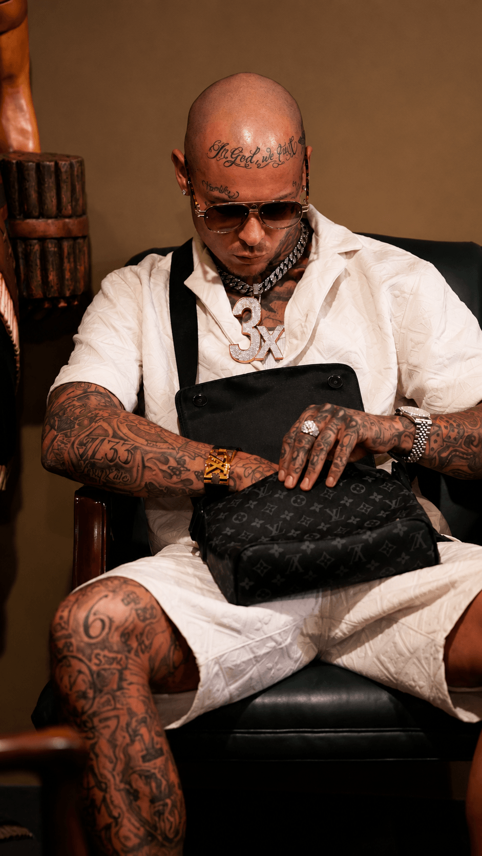 Bald man with tattoos, sunglasses, and white clothing sitting on a black chair, holding a black bag.