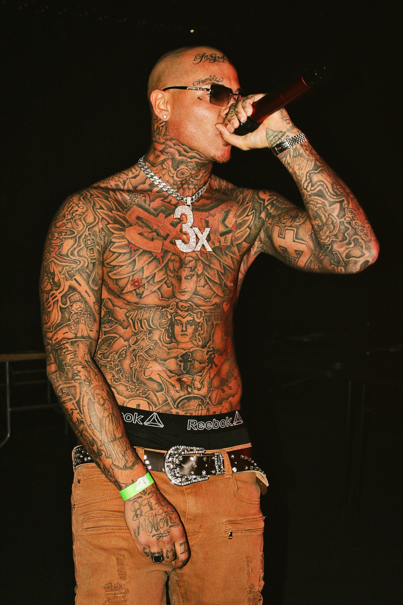 Tattooed man singing into microphone, wearing sunglasses, with a belt and wristbands, against a dark background.