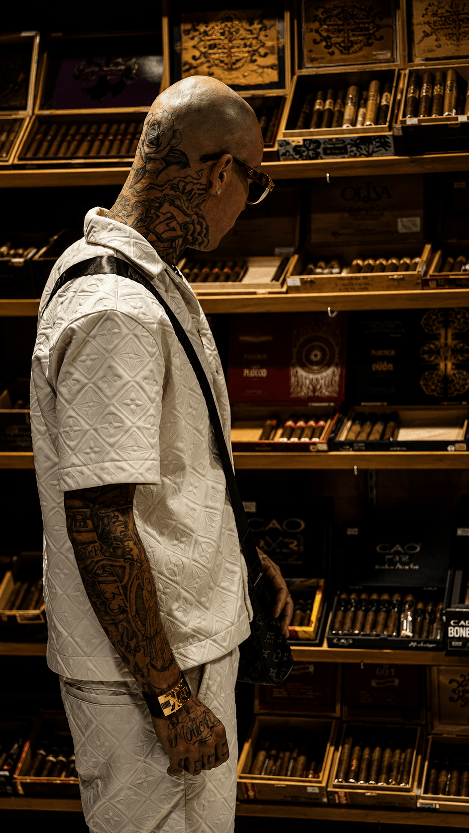 Side view of a tattooed man with a shaved head in a cigar shop, surrounded by shelves of cigars and accessories.