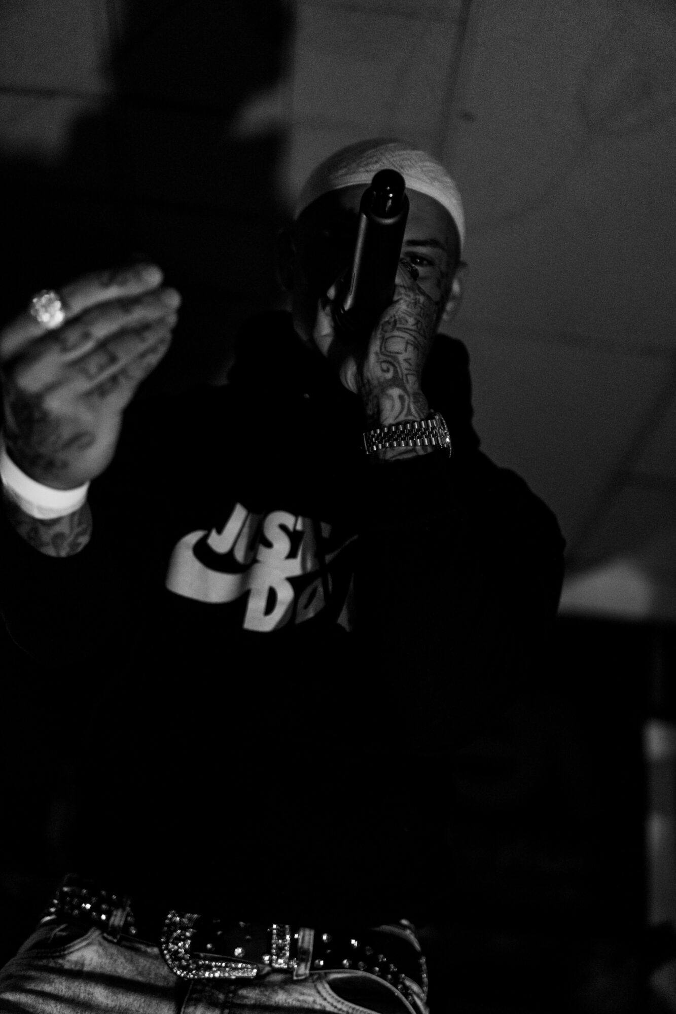 Person wearing a white head covering, holding a microphone, with tattoos and jewelry visible, in a dark setting.
