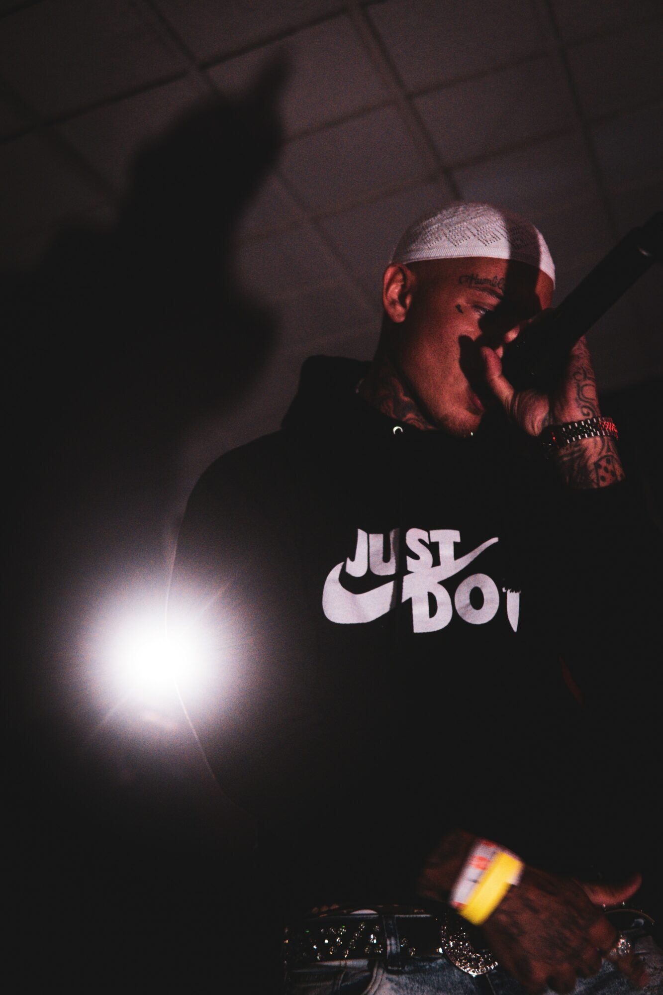Person wearing a hoodie with 'Just Do It' logo, holding a microphone, with a shadow on the ceiling, in a dark setting.