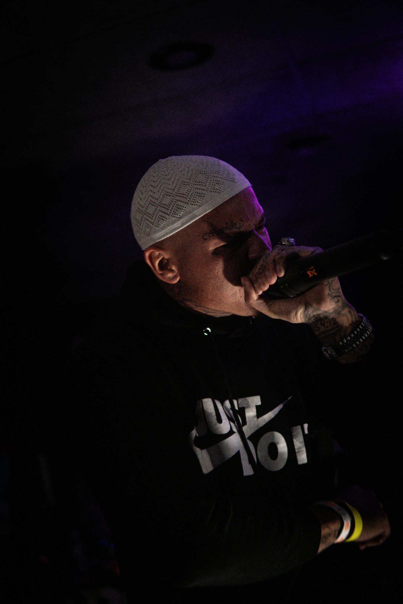 Person wearing a white cap, holding a microphone, in a dark setting with purple lighting.