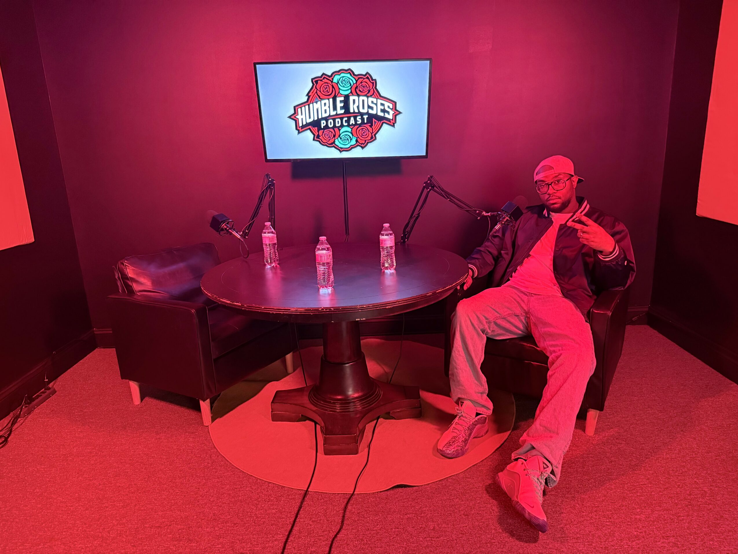 Person sitting on a chair with a microphone, three water bottles on a round table, TV screen displaying 'Double Roses' logo, red lighting