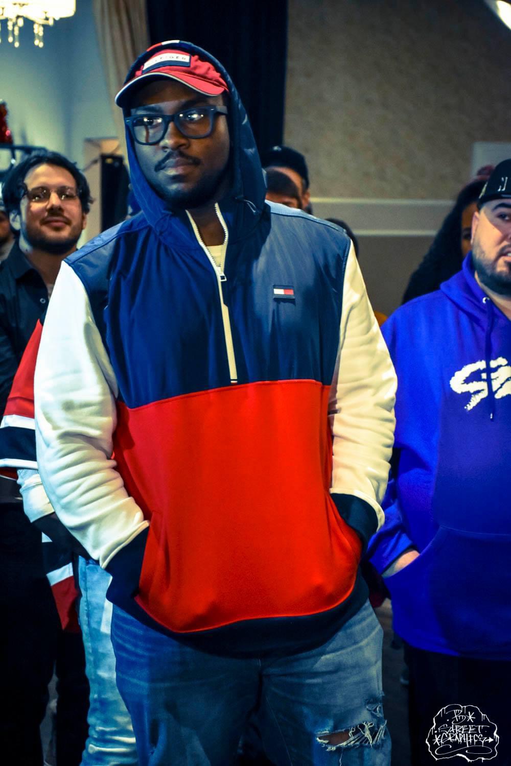Young man wearing glasses, a cap, and a colorful jacket, standing indoors with others in the background.