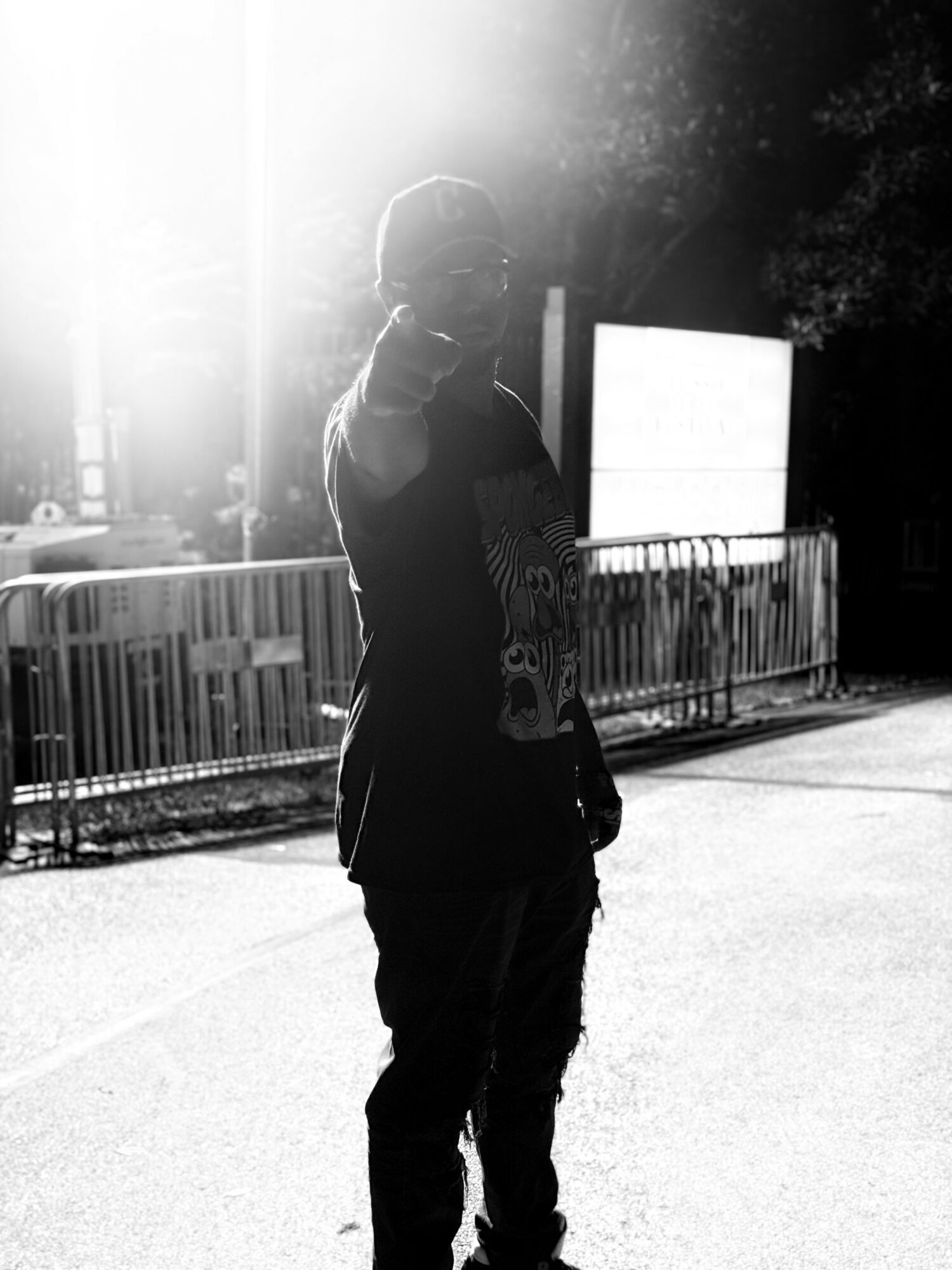 Person standing outdoors near a fence, pointing towards the camera with sunlight behind them, creating a silhouette effect.