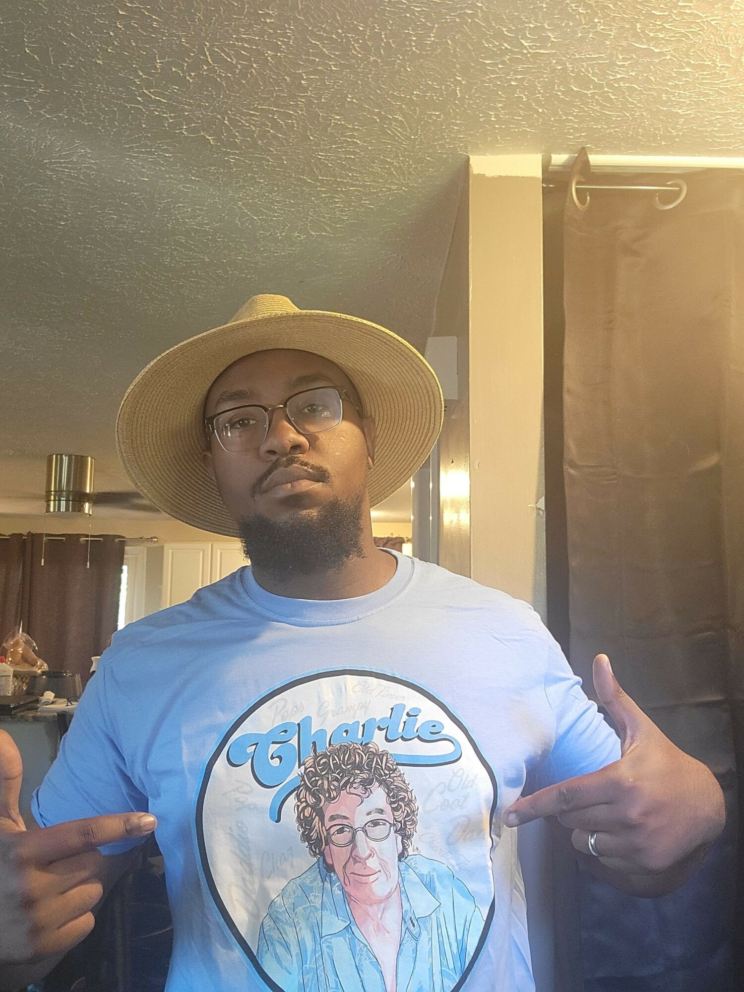Person wearing a wide-brimmed hat and glasses, pointing with both hands, standing indoors with a green wall background.