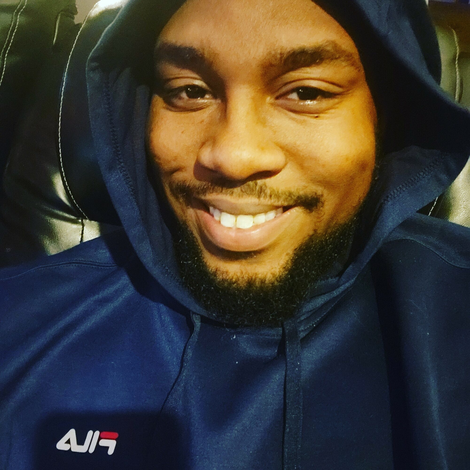 Smiling person wearing a dark hoodie, sitting on a black leather seat, looking at the camera.