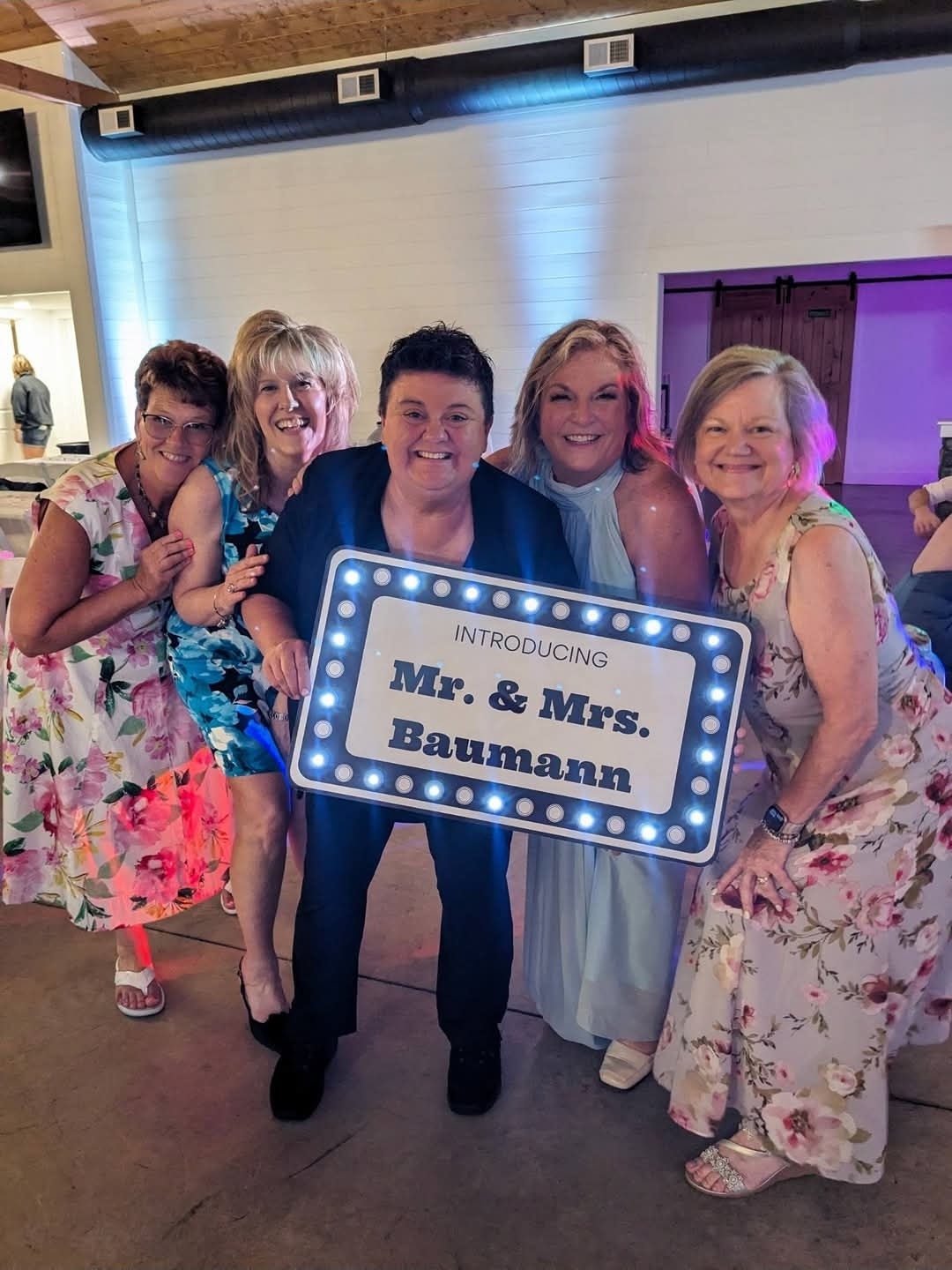 Group of five people holding a sign that says 'Introducing Mr. & Mrs.. Baumann' at a celebration, smiling and dressed in colorful attire.
