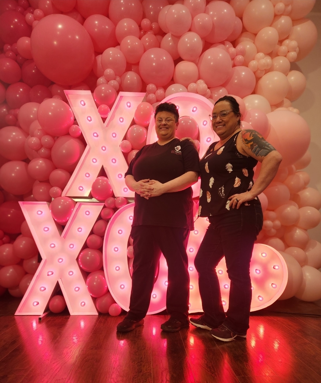 Two people standing in front of a pink balloon backdrop with illuminated letters, smiling at the camera.