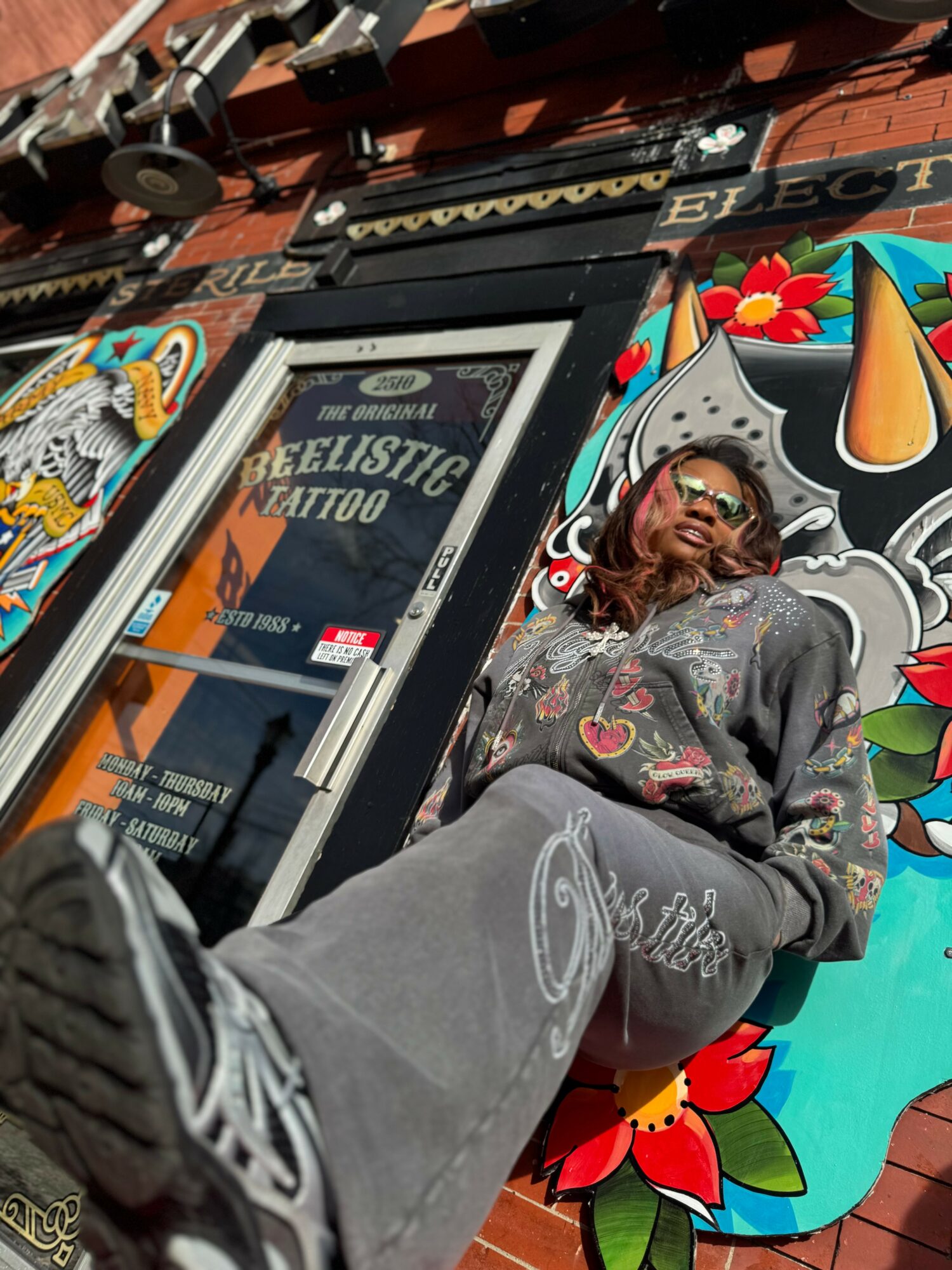 Person with long hair leaning against a colorful mural outside a tattoo shop, wearing a dark hoodie and pants, with graffiti art background.