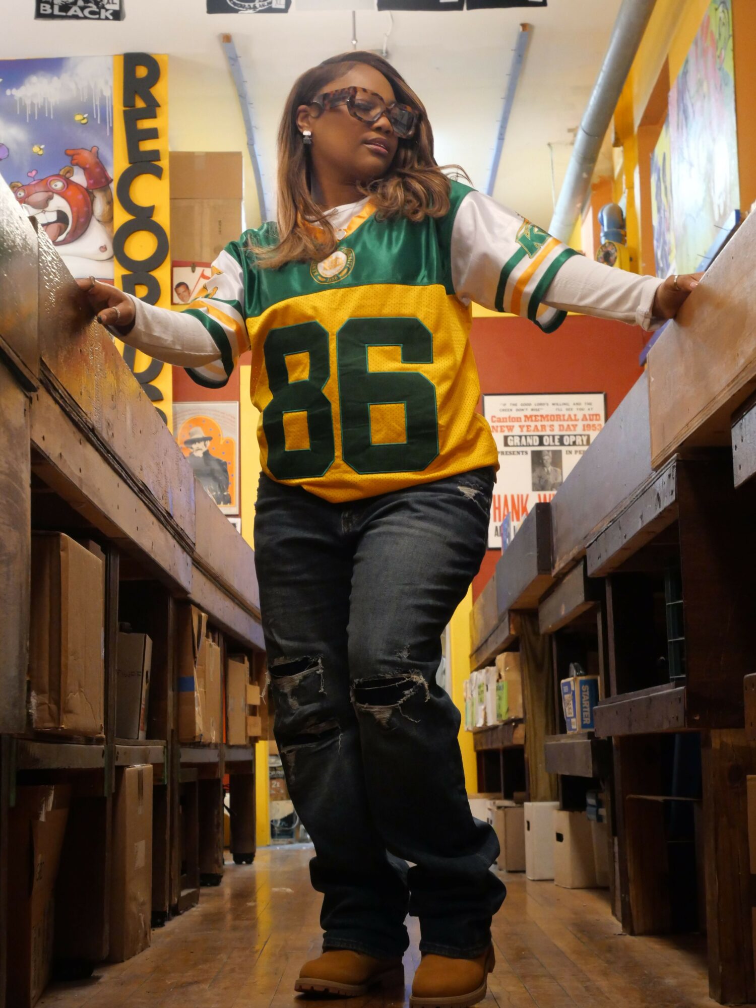 Person with long hair wearing a green and yellow sports jersey, standing in a room with wooden furniture and posters.