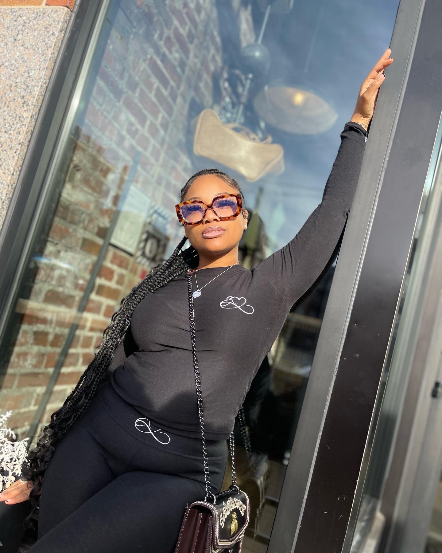 Woman with braided hair and sunglasses standing by glass door, arm raised, brick wall reflection behind her.