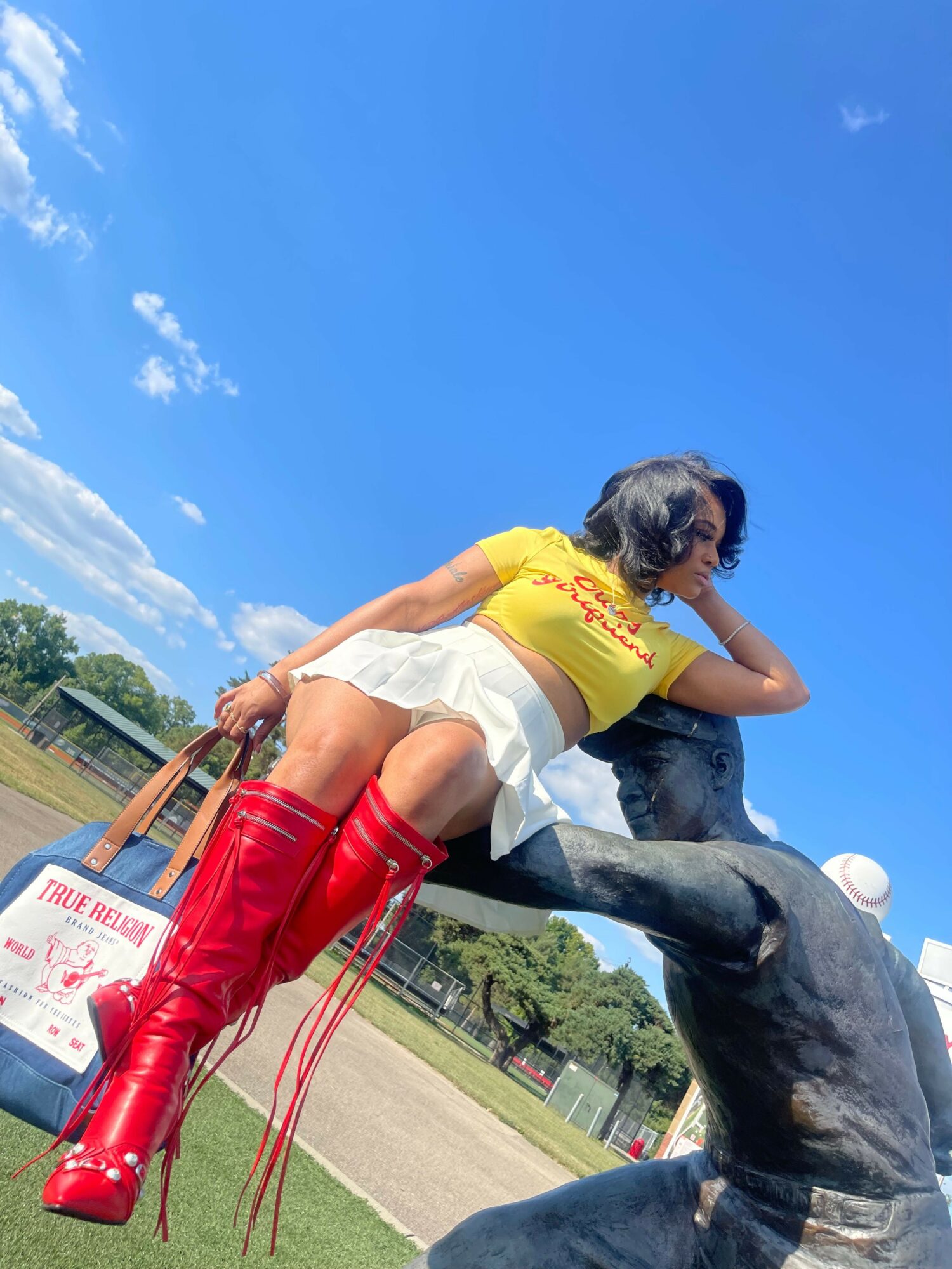 Person wearing red thigh-high boots and a yellow top, sitting on a statue outdoors with a blue sky background.