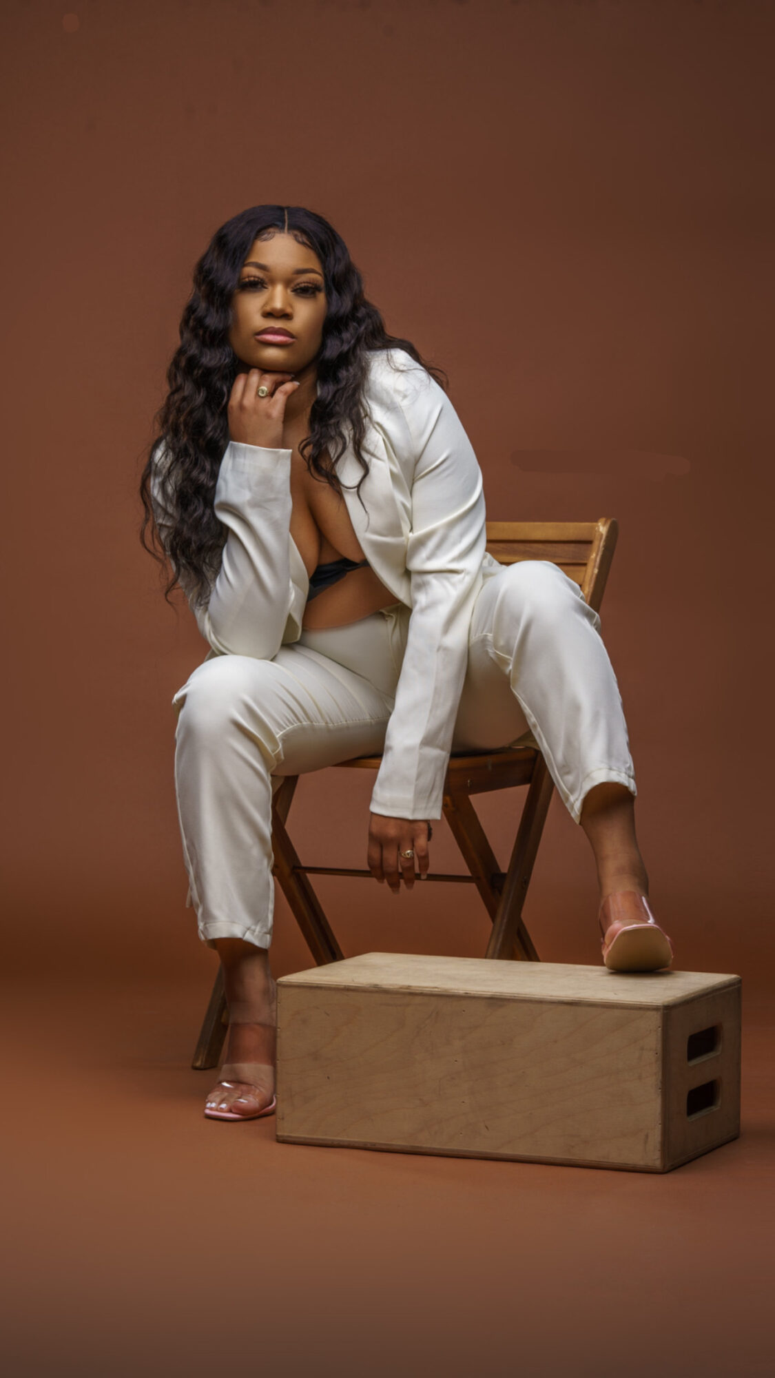 Woman with long wavy hair sitting on a chair, wearing a white outfit, with a brown background.