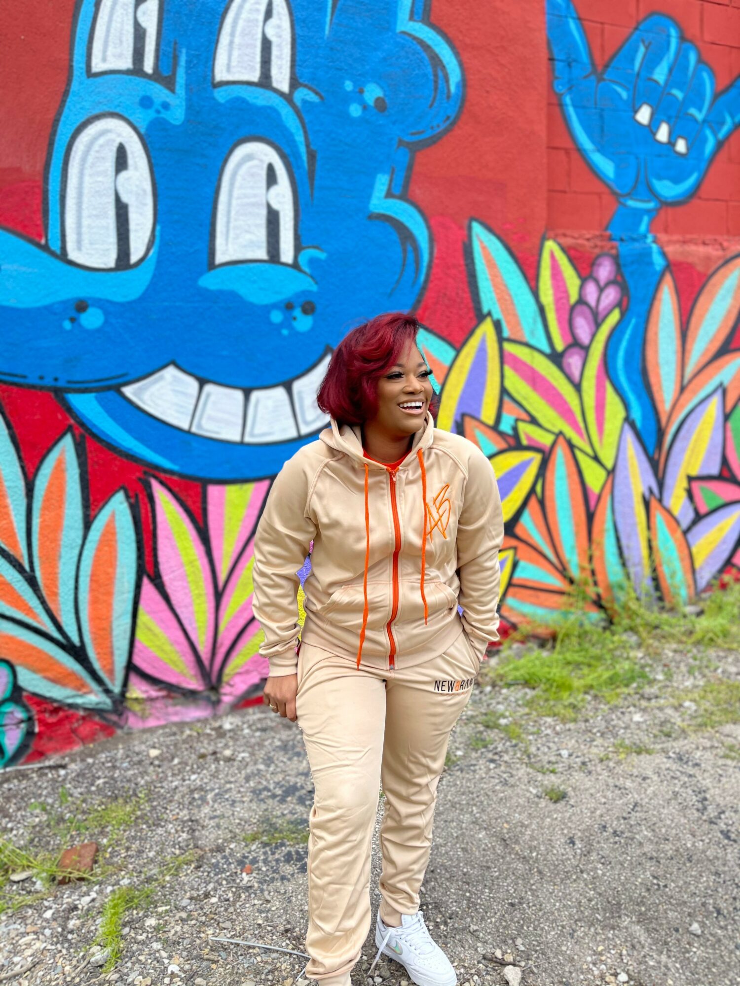 Woman with red hair standing in front of colorful graffiti wall with a blue cartoon character and flowers.