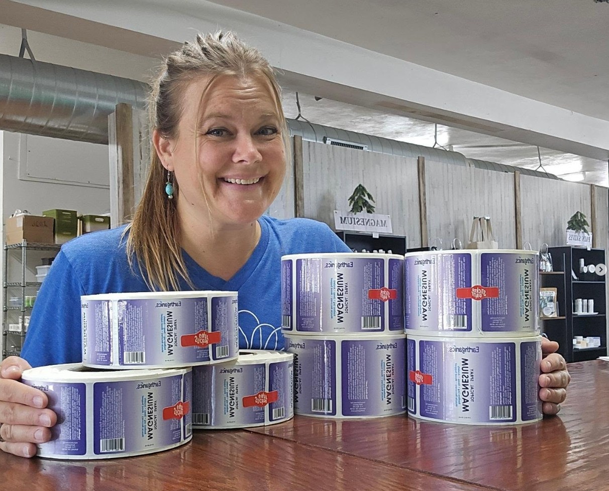 Smiling woman with blonde hair and turquoise earrings displays cans on a wooden table in a store or warehouse.