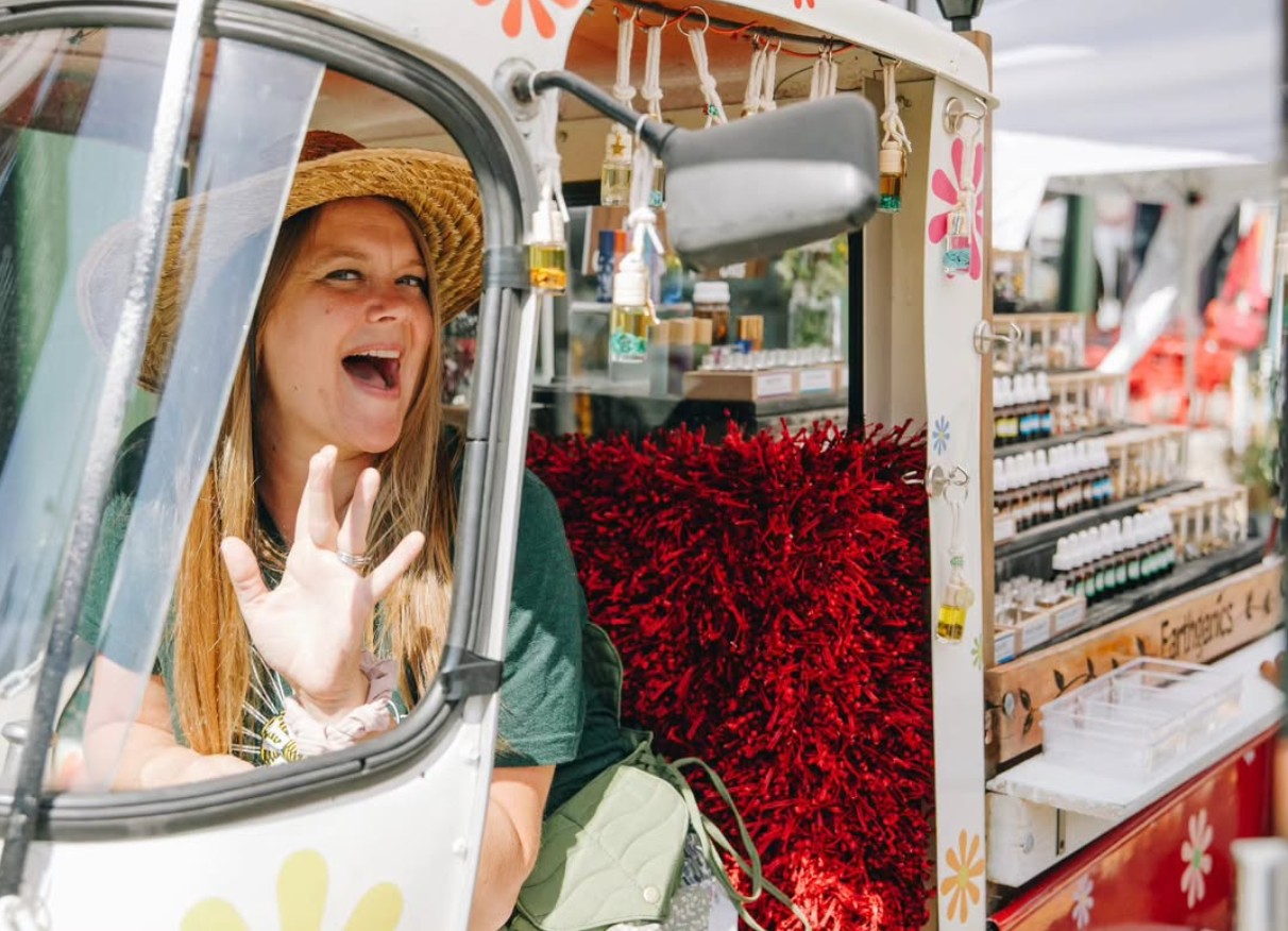 Woman wearing a straw hat smiling and waving from a food truck with red decorations and various supplies.