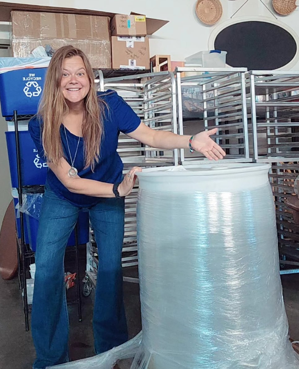 Woman smiling and gesturing next to a large roll of plastic wrap in a warehouse setting.