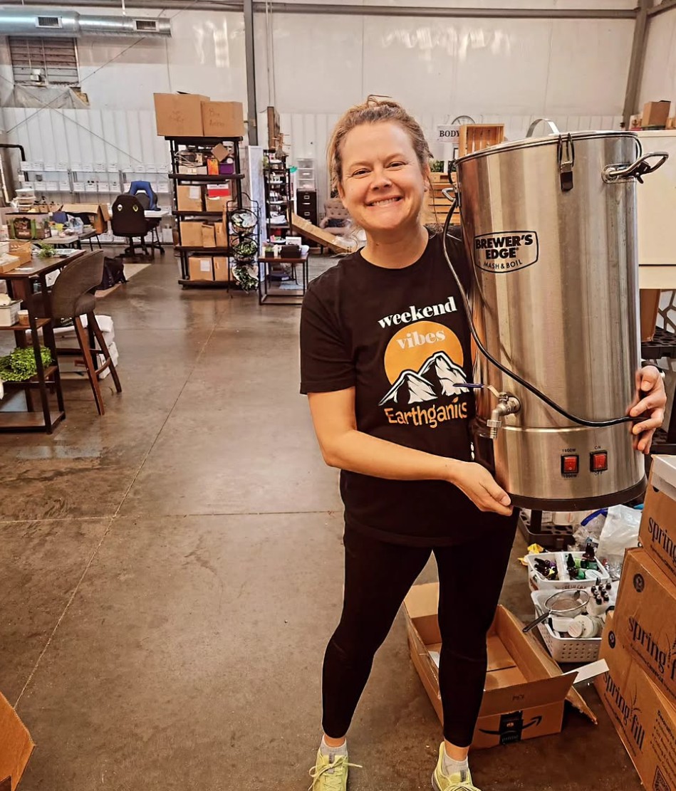 Smiling woman holding a large stainless steel container in a warehouse setting, surrounded by shelves and boxes.